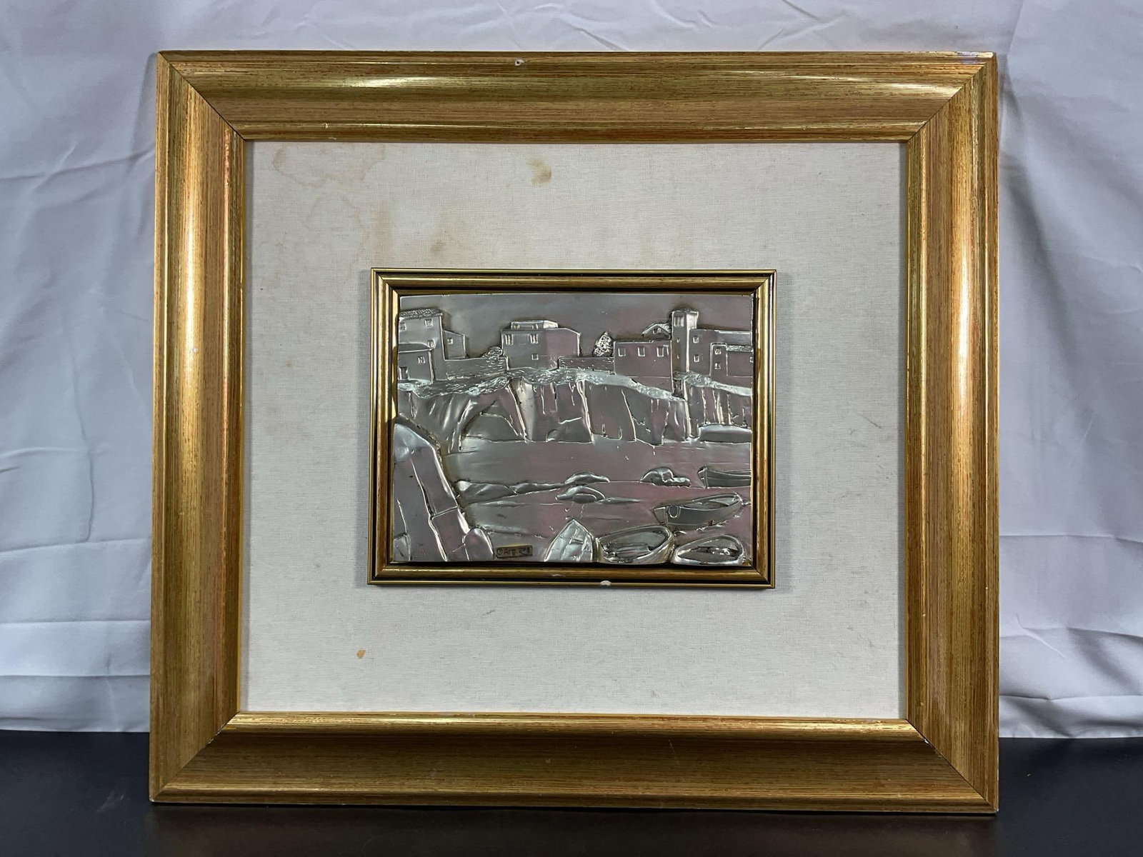 Silver Relief Coastal Scene: A striking metal relief artwork depicting a coastal village perched on cliffs overlooking a body of water. The scene features small boats resting along the shore and a stone bridge connecting the land