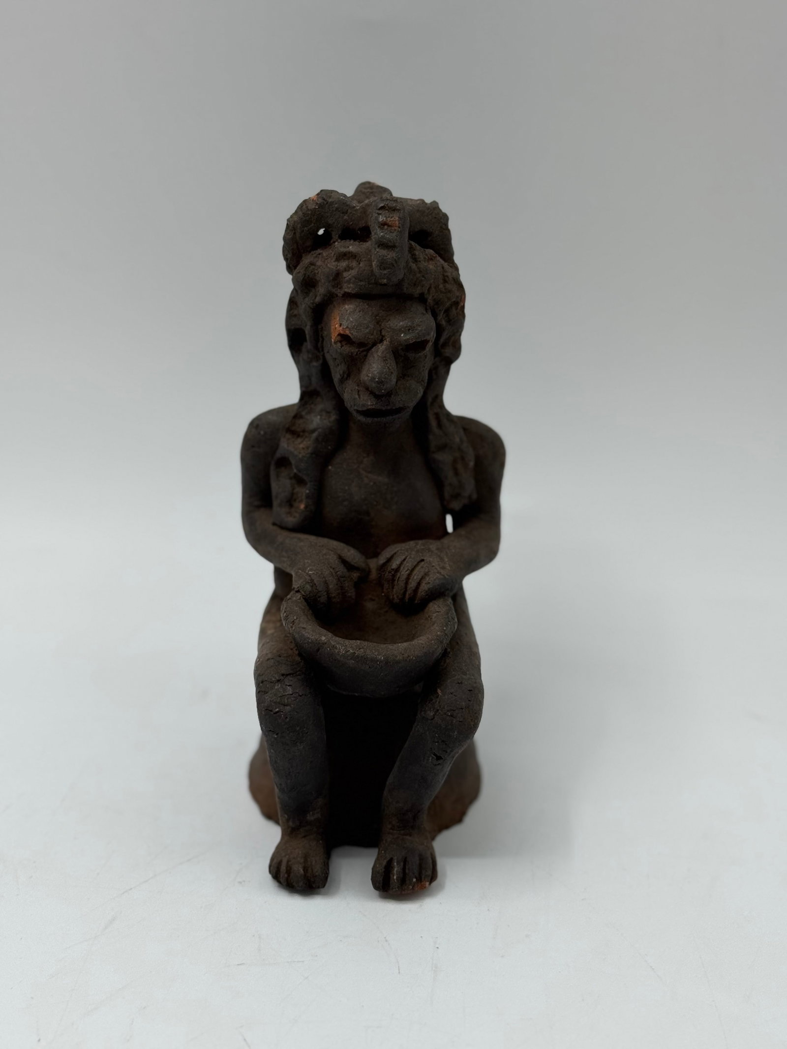 Seated Figure Holding Bowl, Possibly Tribal: A dark-colored sculpture of a seated figure holding a bowl. The figure has detailed hair or headdress and appears to be made of a rough material, possibly clay or wood. Dimensions approximately 6.5 x
