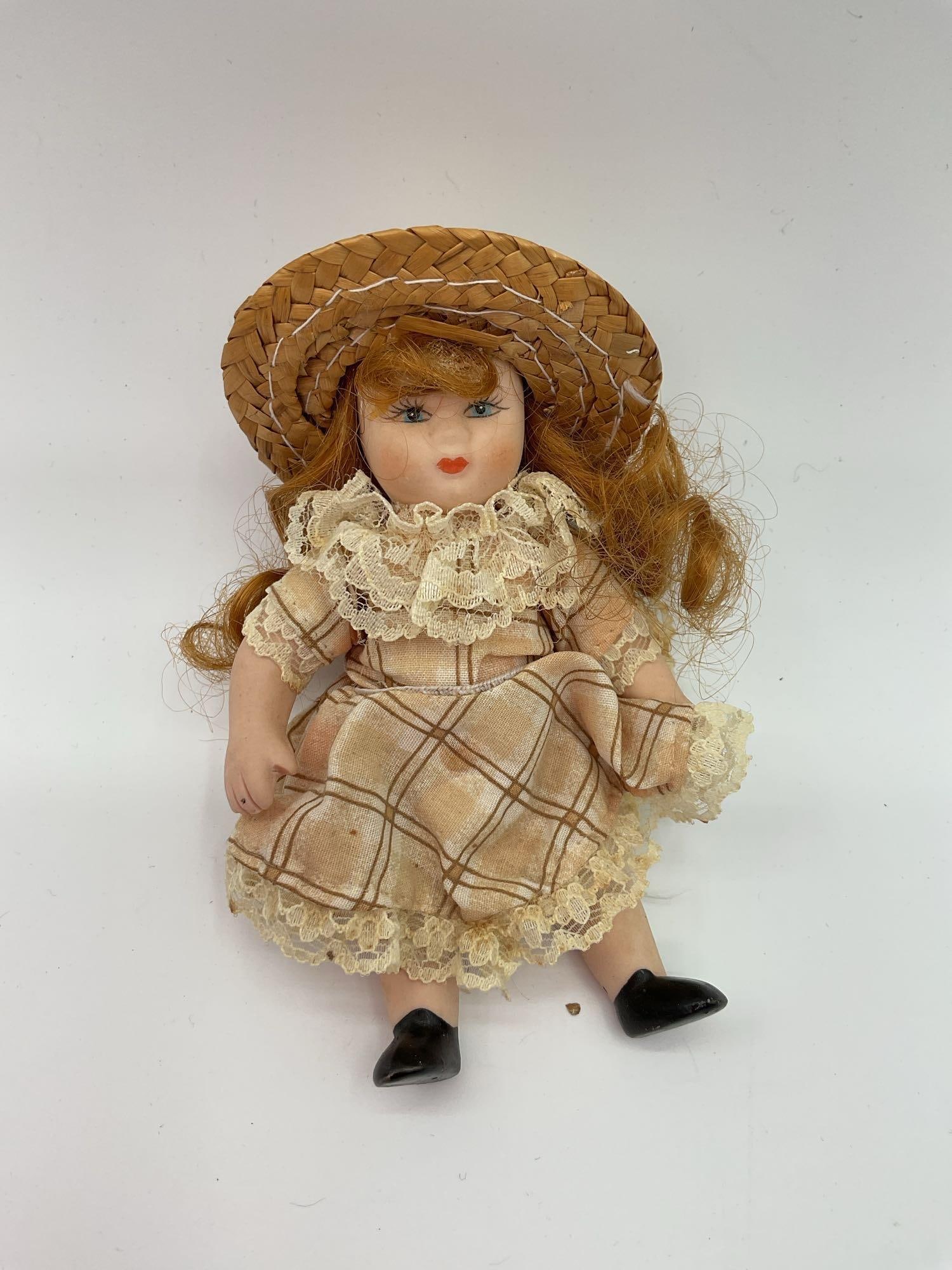 Antique-Style Miniature Porcelain Doll with Straw Hat: This miniature porcelain doll is dressed in a plaid dress with lace trim and a matching straw hat, giving her a rustic, pioneer-era aesthetic. She has red hair, hand-painted facial features with rosy