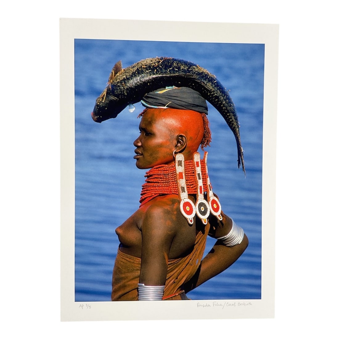 "Dinka Woman with Fish, South Sudan" by Angela Fisher (Australia, B.1947) and Carol Beckwith (USA,: This vibrant color photograph captures a Dinka woman in South Sudan, distinguished by traditional red pigment on her skin and elaborate beaded necklaces. She is depicted in profile against a deep blue