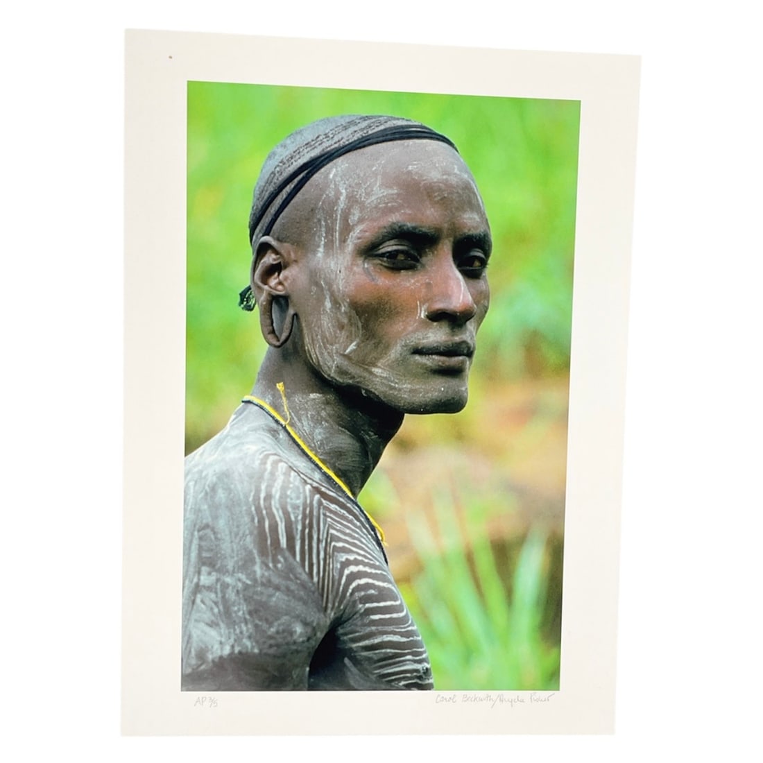 "Maasai Warrior, Kenya" by Carol Beckwith (USA, B.1945) and Angela Fisher (Australia, B.1947) Artist: This color photograph captures a ceremonial portrait of a Maasai warrior in Kenya. The subject features traditional white chalk body ornamentation in a chevron pattern across his torso and delicate fa