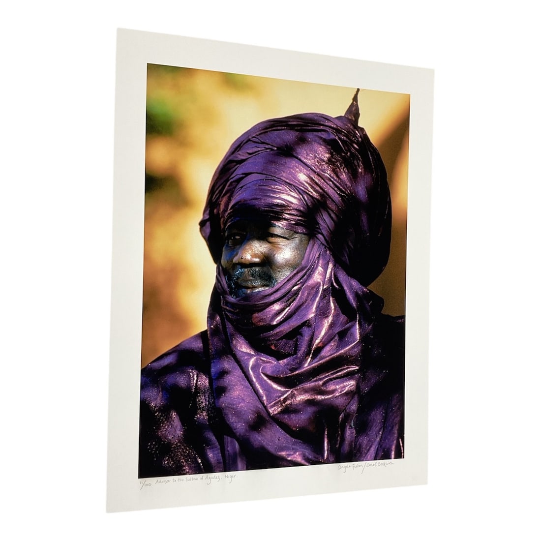 "Dinka Woman with Fish, South Sudan" by Angela Fisher (Australia, B.1947) and Carol Beckwith (USA,: This color photograph captures a Dinka woman in South Sudan, balancing a large fish on her head against a deep blue water background. The subject is adorned with traditional beaded necklaces and red p