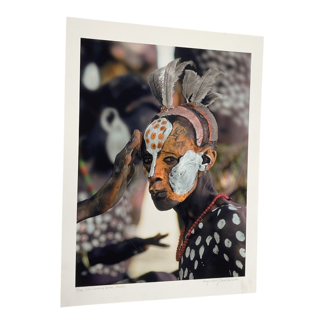 "Surma Boy with White Feathers, Ethiopia" by Carol Beckwith (USA, B.1945) and Angela Fisher: This color photograph depicts a young Surma boy from the Omo Valley in Ethiopia. He is shown with a traditional headdress made of large white feathers and a red tassel, along with intricate white chal