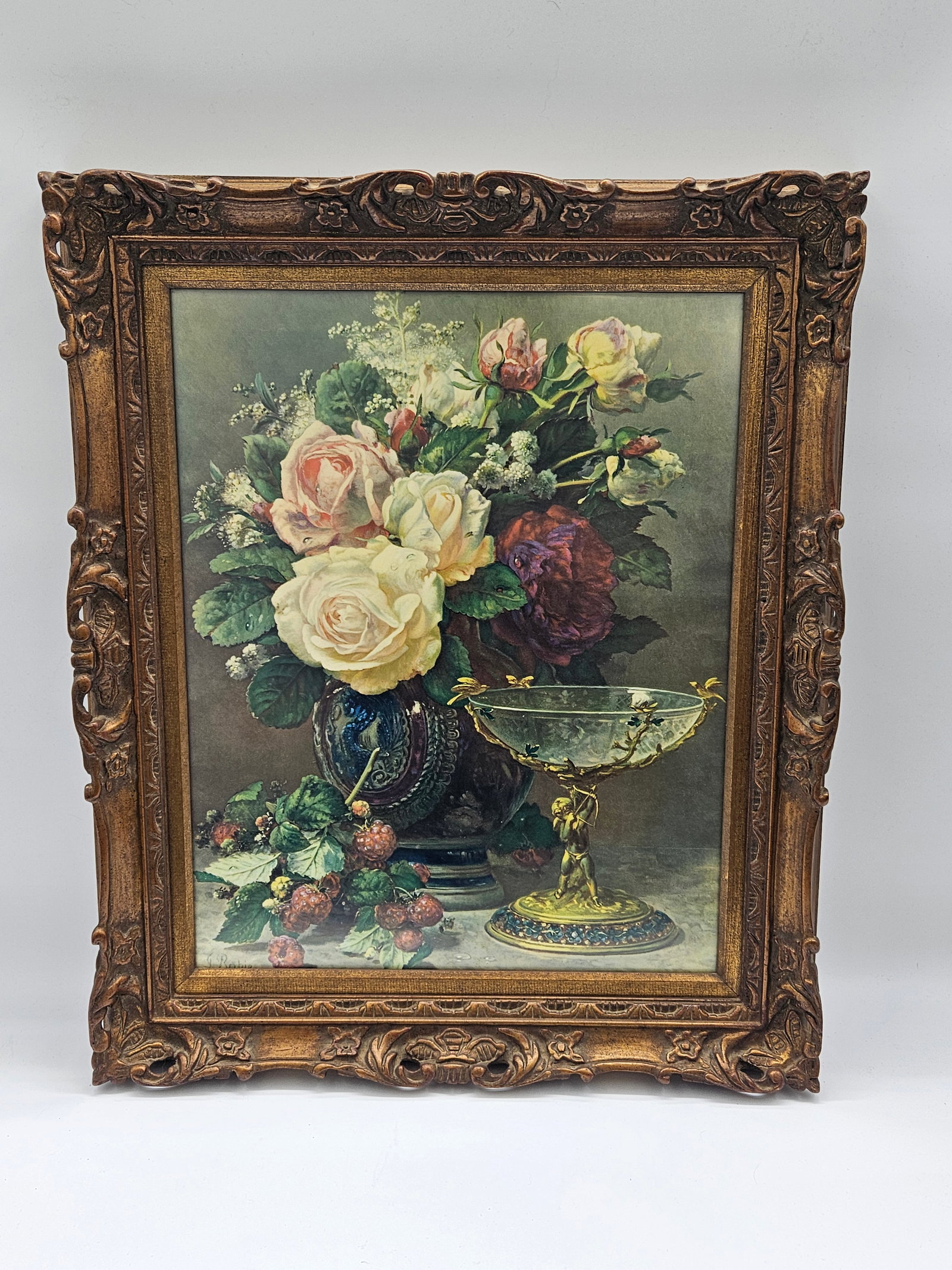 Jean Baptiste G Robie - Cupids Goblet: A framed print depicting a floral still life with roses and other flowers, alongside a decorative vase and mirror. The print is signed 'Robie'. Presented in an ornate gold-tone frame. 25x21in