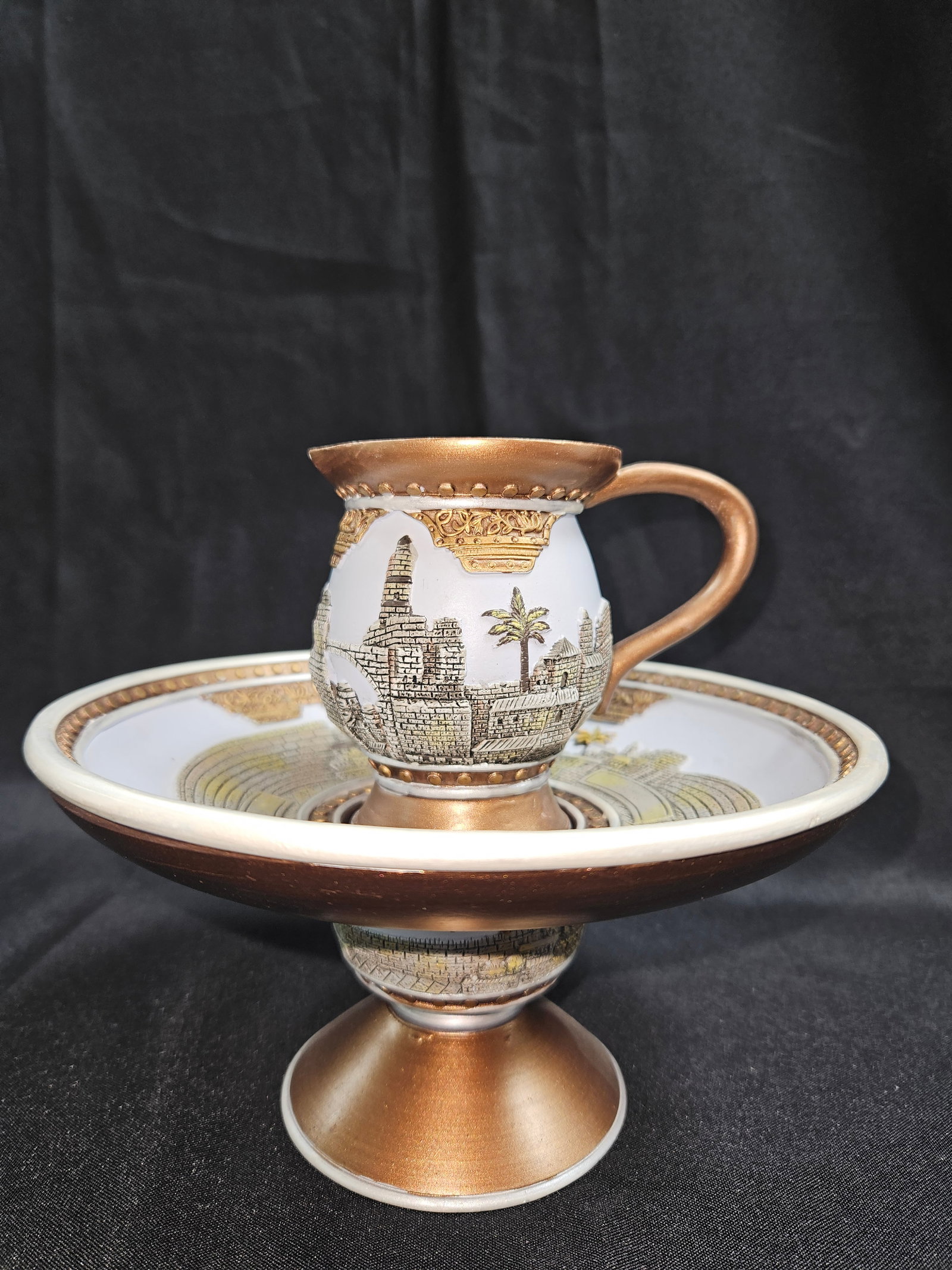 Ceramic Cup and Footed Bowl Set with Cityscape Design: Small ceramic pitcher set atop a matching pedestal bowl, featuring intricate scenic designs highlighted with gold and bronze tones. Likely decorative, not utilitarian.