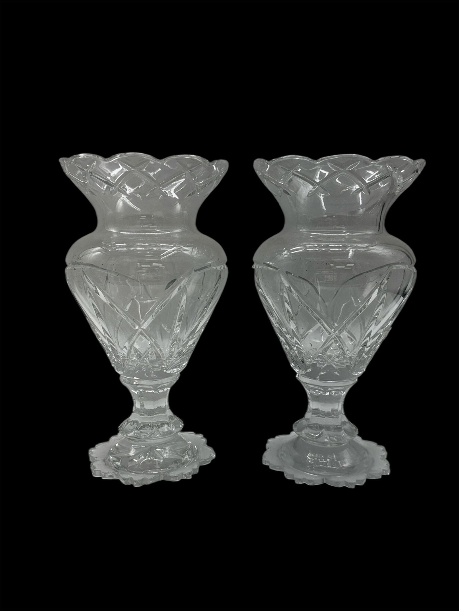 Pair of Cut Glass Pedestal Vases: A pair of clear cut glass pedestal vases featuring flared scalloped rims, faceted bodies, and decorative footed bases. Each vase displays intricate geometric and starburst cutting that enhances brilli