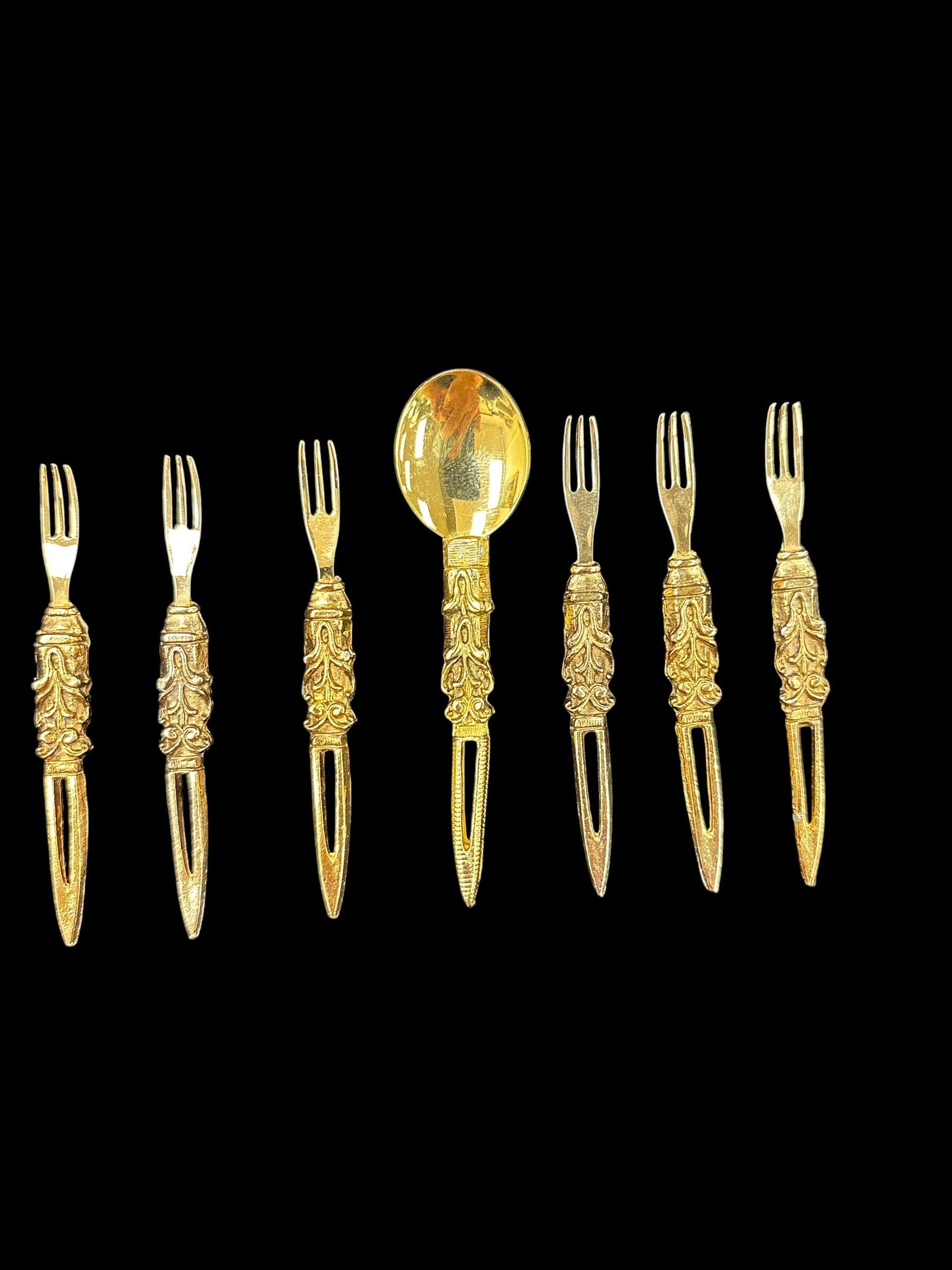 Set of Seven Gold Tone Hors d’Oeuvre Forks with Spoon: A decorative set of seven small serving utensils, including six forks and one spoon, featuring ornate handles with detailed relief designs. The pieces are finished in a gold tone and appear to be cons