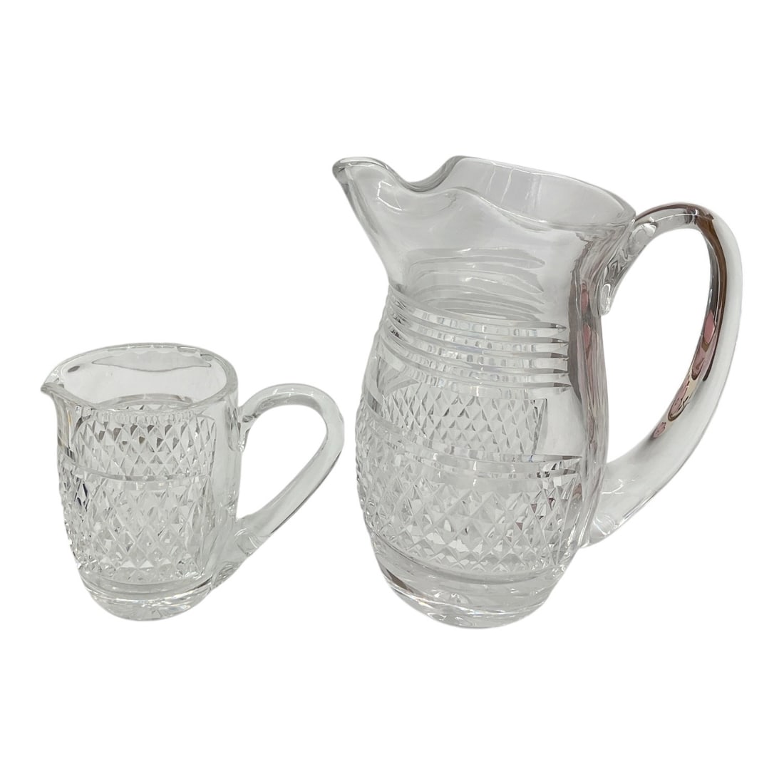 Pair of Cut Glass Pitchers: Decorative pair of clear cut glass pitchers featuring diamond and paneled cut decoration with applied handles and shaped pouring spouts. Includes one larger pitcher and one smaller cream pitcher. Eleg