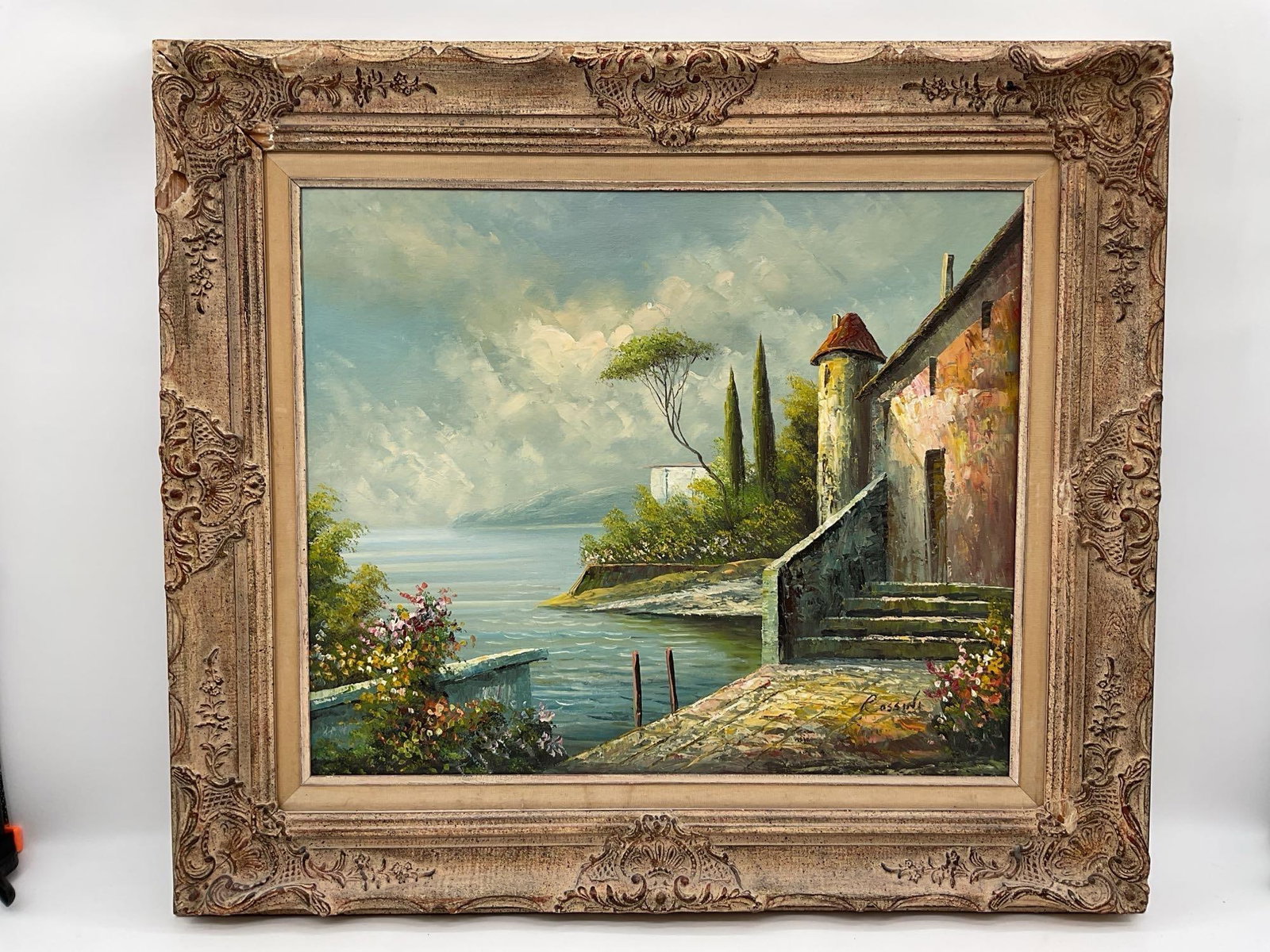 Romano Rossini (Italian, 1886-1951) Oil on Canvas: A serene landscape painting by Italian artist Romano Rossini depicting a quiet villa by the lake, featuring a stone path leading to the water, flowering bushes, and a distant view of rolling hill