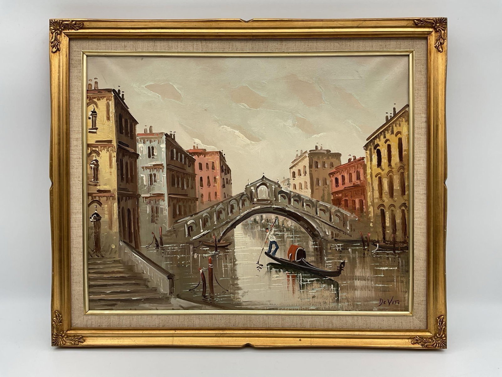 Antonio DeVity (Italian 1901-1993) Oil on Canvas: A classic mid-century oil painting of a gondolier navigating a canal beneath the iconic Rialto Bridge in Venice, Italy. Lined with pastel-colored historic buildings and softly textured brushstrokes,