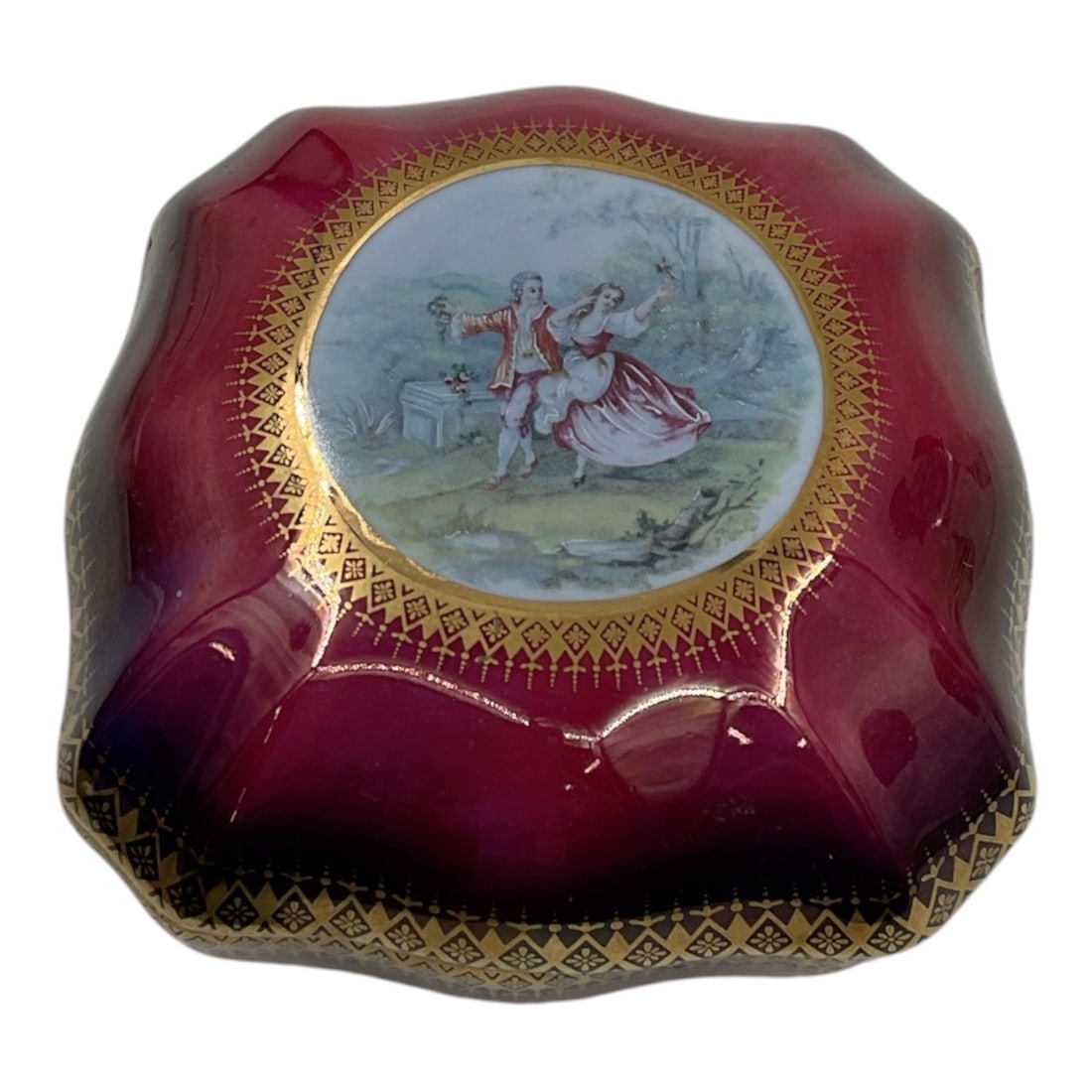 Porcelain Trinket Box with Courting Scene Decoration: Decorative porcelain trinket box and cover featuring a deep cranberry ground with gilt geometric border decoration. The lid is centered with a transferdecorated courting scene depicting an 18thcentury
