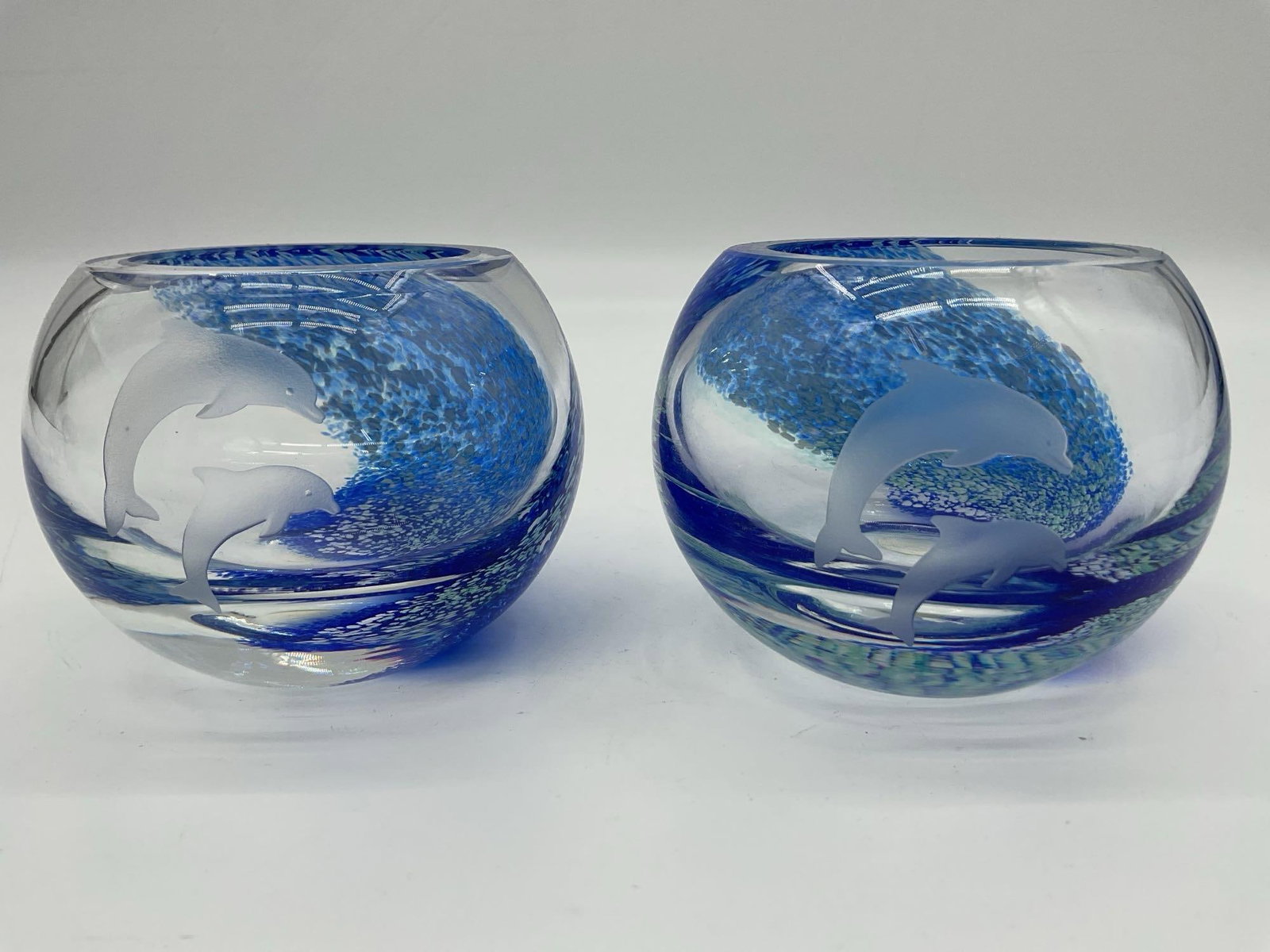 Pair of Lenox HandBlown Dolphin Votive Holders – Made in Scotland: Set of two round art glass votive holders by Lenox, crafted in Scotland. Each features a swirl of cobalt and aqua speckling encased in clear glass, with one piece showcasing an etched dolphin motif. E