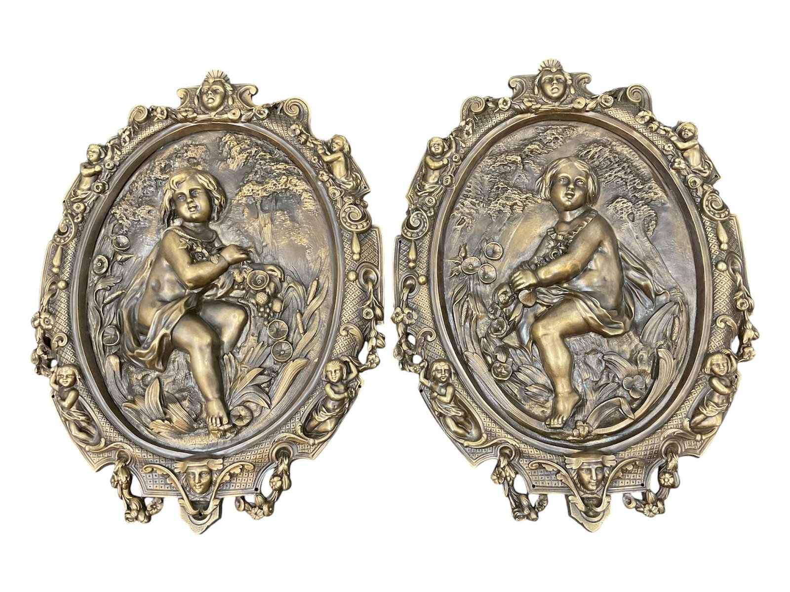Pair of Neoclassical Putti Relief Wall Plaques (Antique) Brass or Bronze: This matching pair of antique oval wall plaques features high-relief sculptural depictions of winged putti or cherubs nestled within a pastoral landscape. Each figure is shown seated among lush foliag