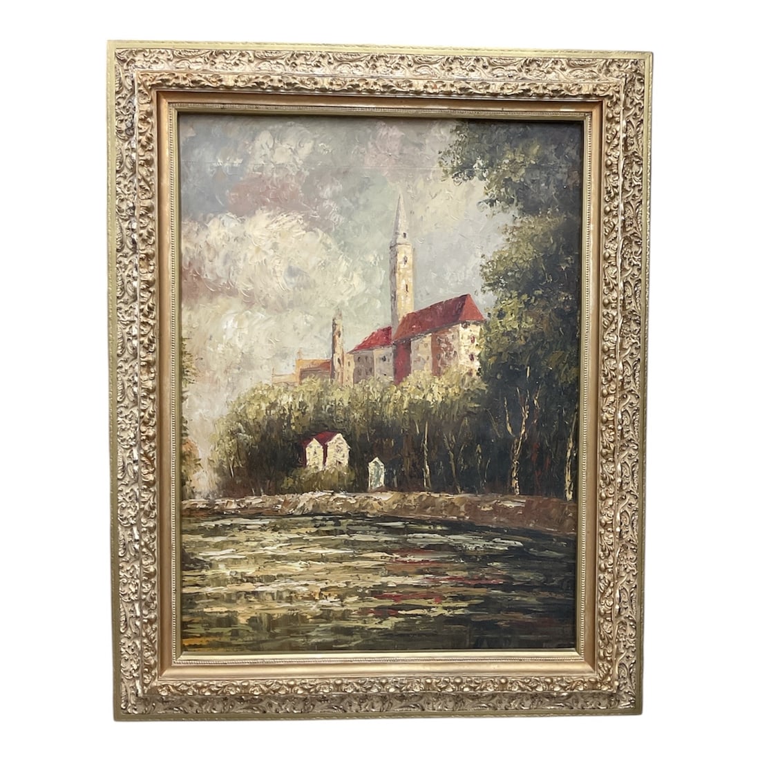 European Village Landscape Oil Painting Signed: A large framed oil painting depicting a picturesque European village scene with a church featuring a tall steeple and red roof, set among trees and overlooking a reflective body of water. The composit