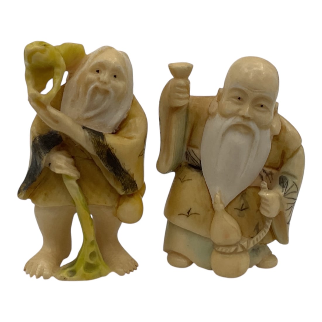 Pair of Asian Carved Elder Immortal Figurines: A pair of miniature Asian carved-style figurines depicting elderly male immortals or sages with long flowing beards and traditional robes. One figure is portrayed holding a staff beside a naturalistic