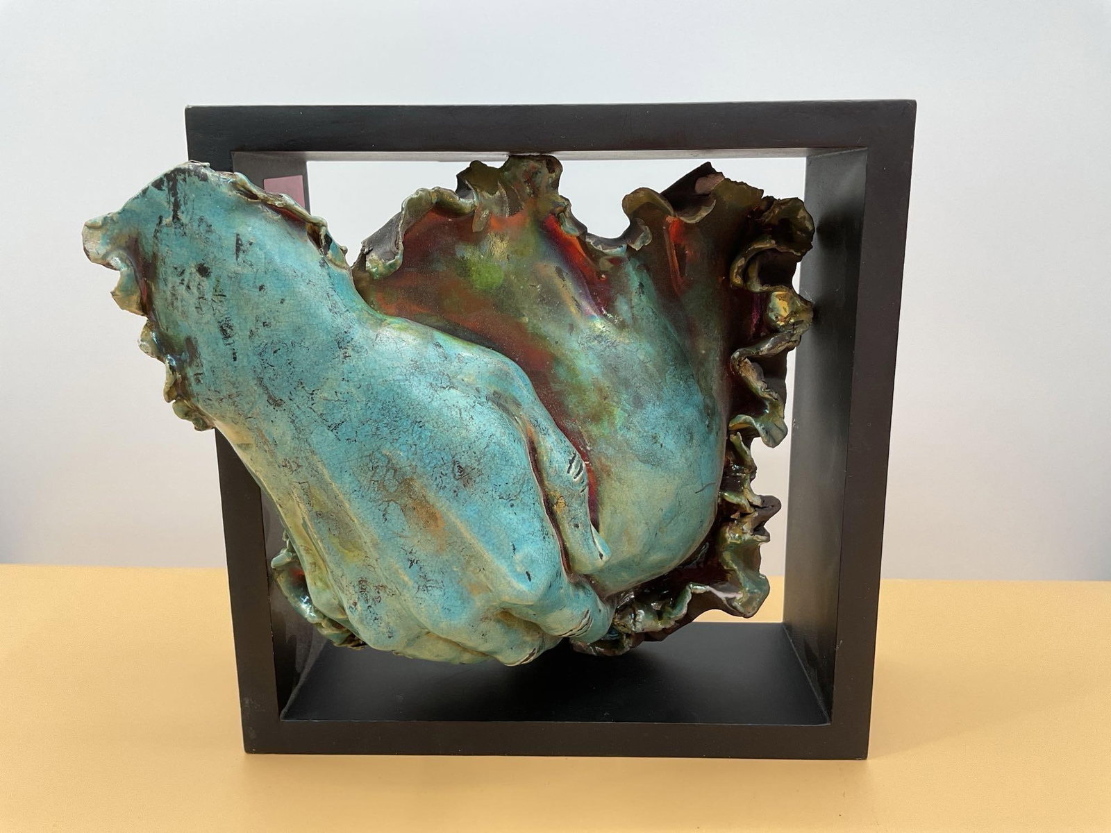 Raku Ceramic Sculpture of Clasped Hands - Signed: This expressive ceramic sculpture features a pair of clasped hands resting on an organic, ruffled base. Finished in a rich, iridescent raku glaze with turquoise and metallic tones, the surface display