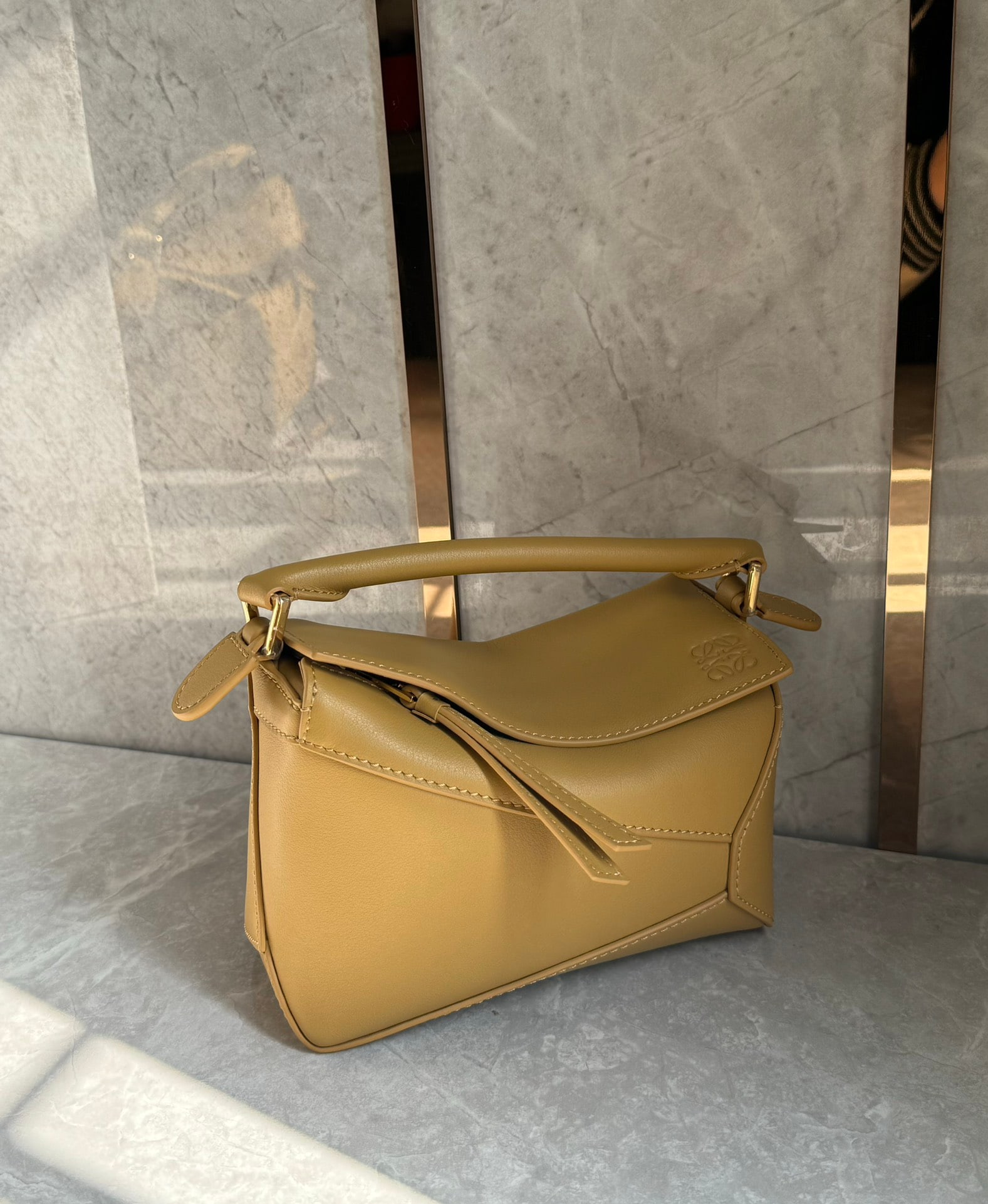 LOEWE: The LOEWE Puzzle mini bag in desert yellow features an adjustable shoulder strap and is crafted from classic pebbled cowhide leather, giving it a luxurious feel. With ample capacity, the Puzzle is