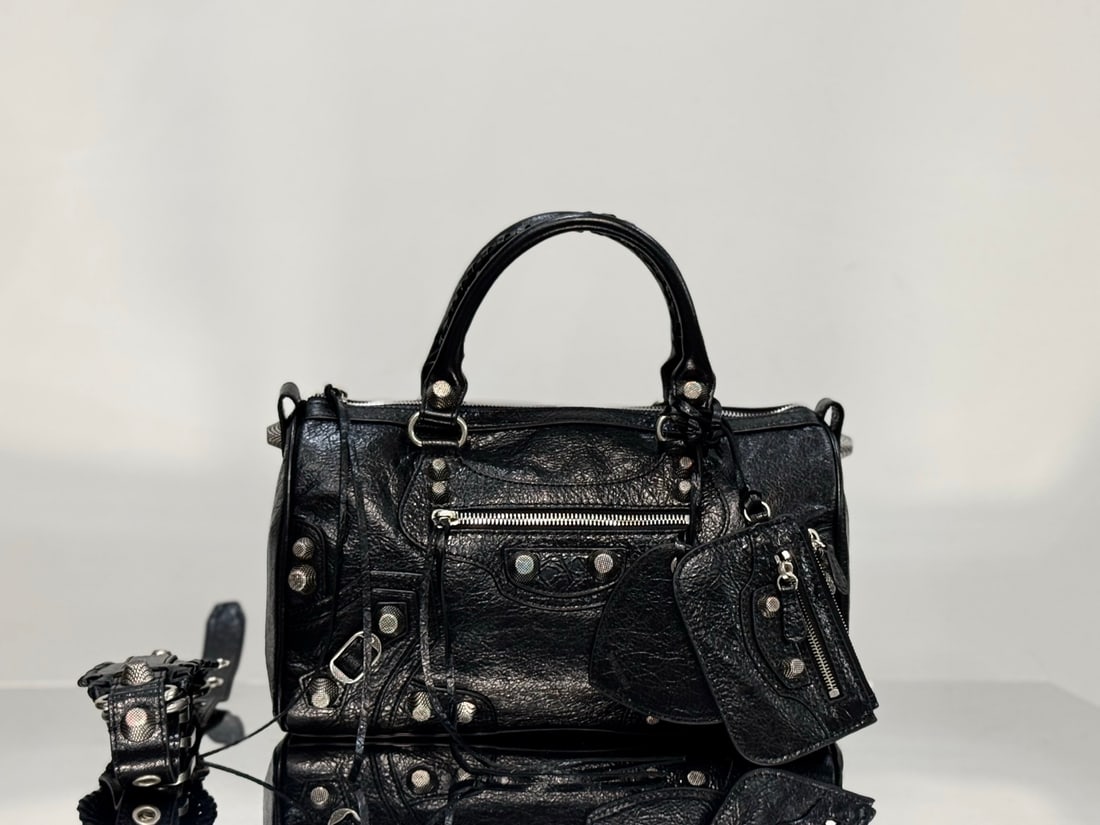 Balenciaga Le Cagole: This Balenciaga Le Cagole duffle bag is a mini travel bag made of black fine-grained lambskin leather with silver hardware. It retains the classic motorcycle bag silhouette, featuring a unique and pra