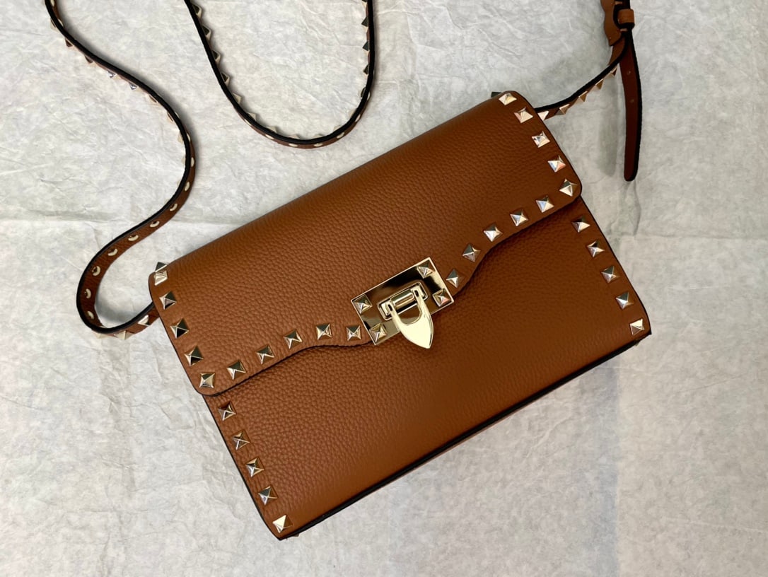 Valentino: Brown Valentino messenger bag with tumbled gold studs and a simple design. Features an adjustable shoulder strap and one interior slip pocket. Leather: Plain tumbled cowhide. Size: 22.5 x 5 x 16 cm.