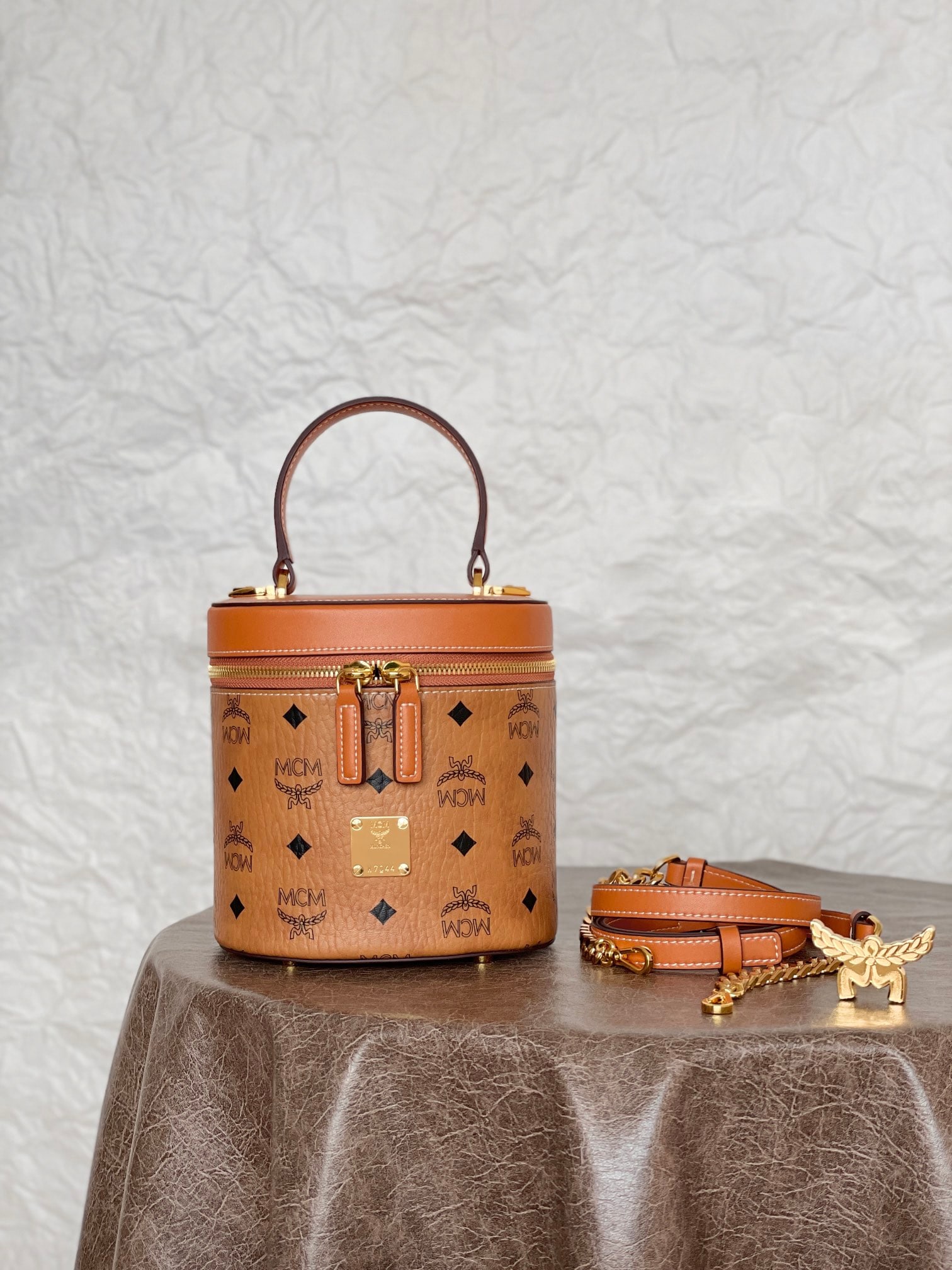 MCM AREN VISETOS Lucky Bucket Bag: MCM AREN VISETOS Prosperity Bucket Bag in Cognac Monogram. This spacious cylindrical crossbody bag features a geometric design adorned with the classic Visetos print and a vintage gold-tone logo