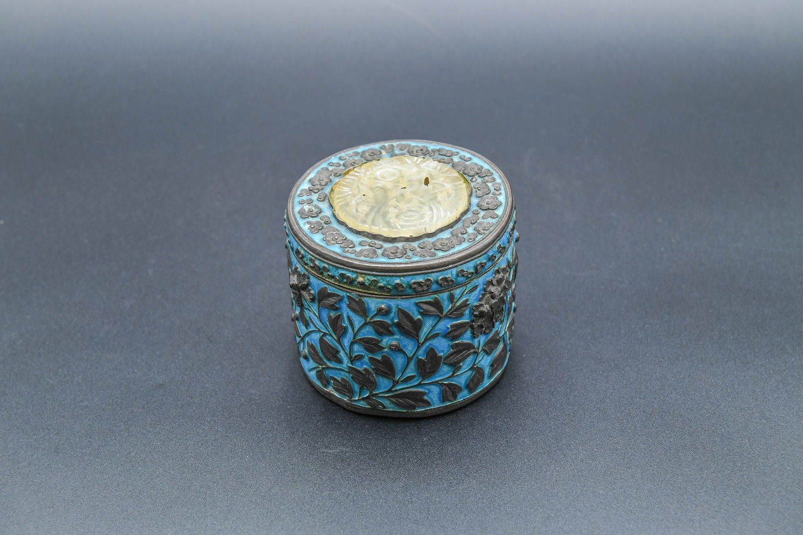 Antique Chinese Cloisonne Enamel Lidded Round Box: Antique Chinese Cloisonne Enamel Lidded Round Box. The box features a vibrant turquoise blue enamel background with dark, raised floral and leaf motifs. The lid is topped with a circular inset of a ca