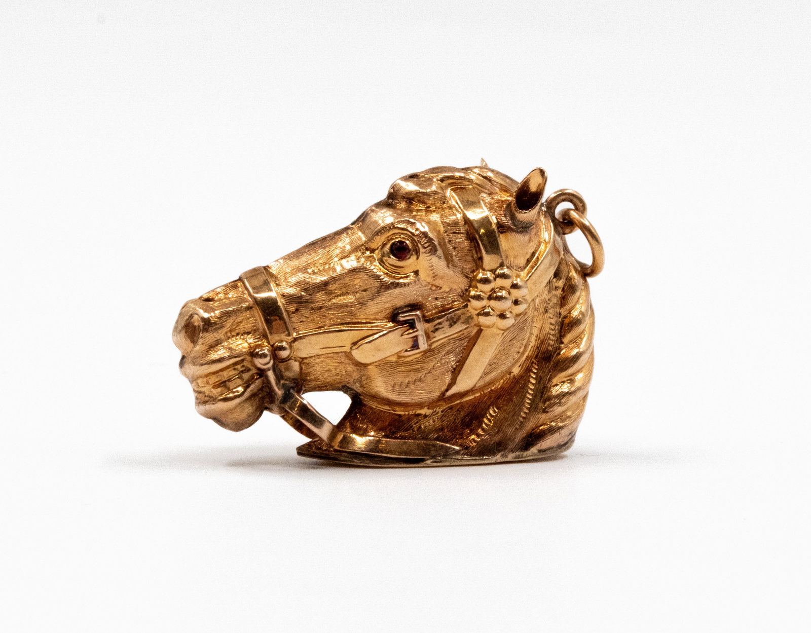 A 14k gold horse head shaped pendant: A 14k gold horse head shaped pendant. Modelled as a bridled horse's head with textured mane and details, set with a red stone eye and suspension loop. Total weight: 8,7g
