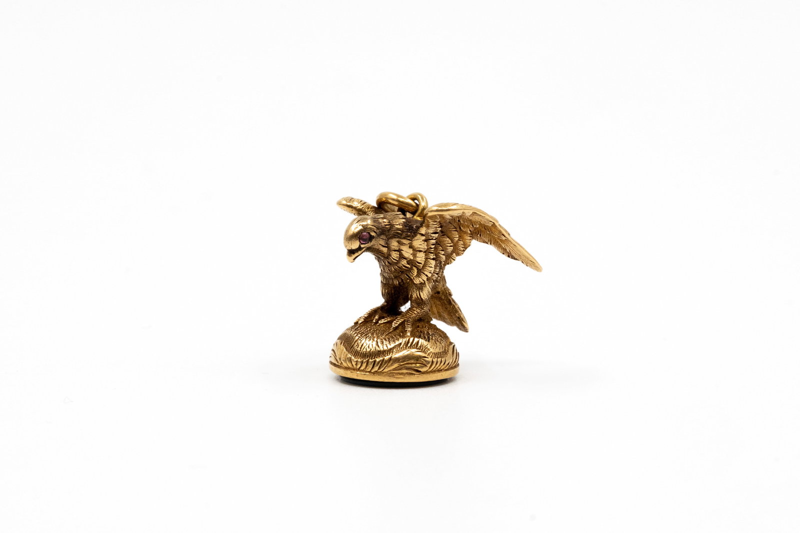 A 18k gold bird seal pendant: A 18k gold bird seal pendant. Modelled as a bird of prey with textured feathers, set with a red stone eye, standing on a circular seal base engraved with a monogram. Total weight: 9g H: 1,7 - W: 1,5 c