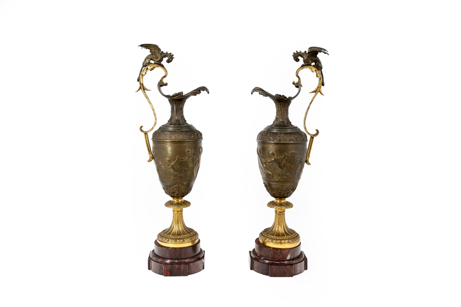 A pair of large and antique gilt and patinated bronze ewer jugs: A pair of large and antique gilt and patinated bronze ewer jugs decorated with bas-reliëf scenes and figure of a dragon on top. Mounted on marble bases. Circa 1900. Condition: both marble bases have 