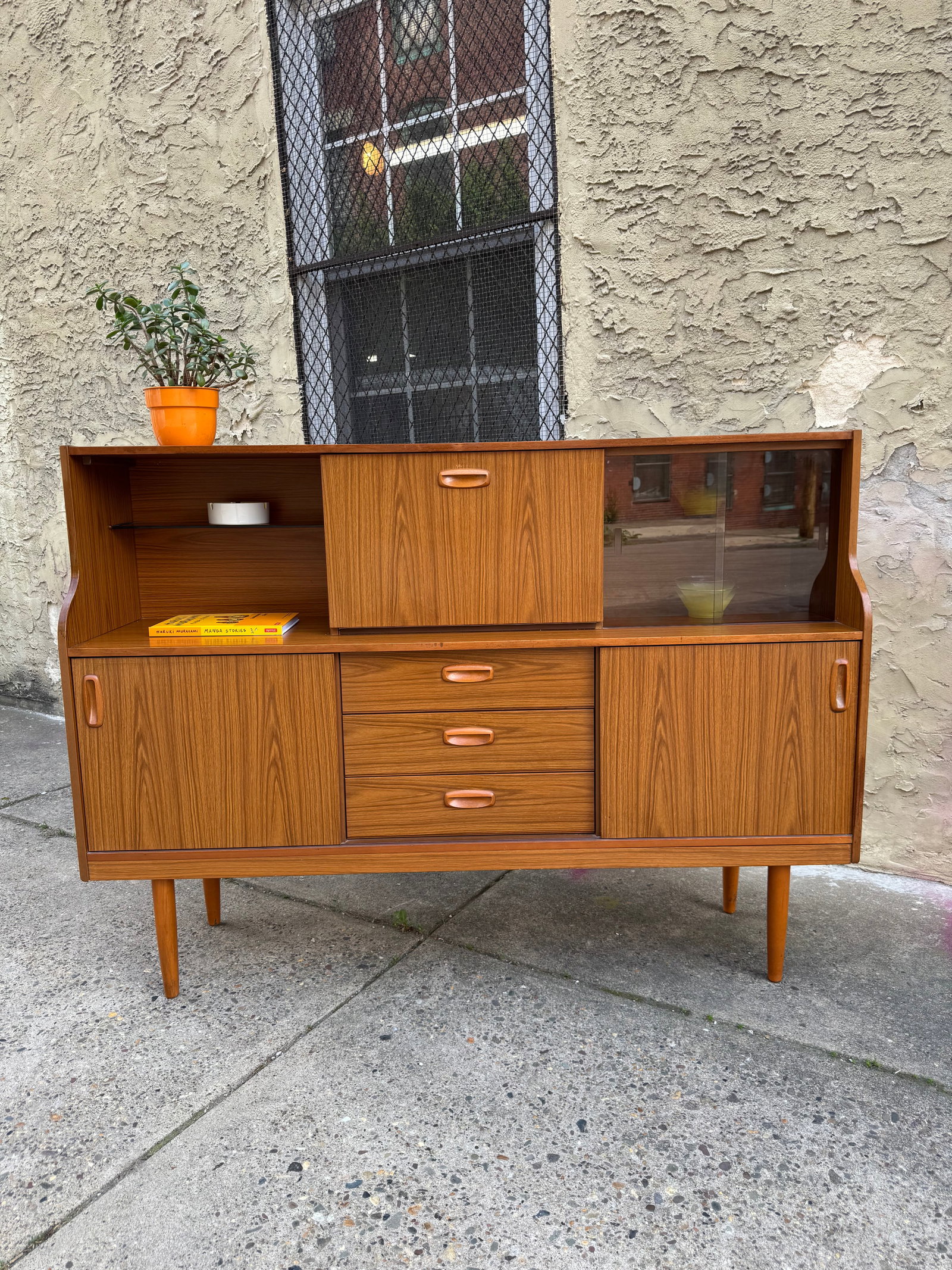 Schreiber Mid Century Teak Credenza, Drop Front Bar Cabinet: Mid-century Schreiber highboard cocktail cabinet from the 1960s–70s, featuring teak-effect veneer with signature recessed pulls and tapered legs. The upper section includes a glass-front display and