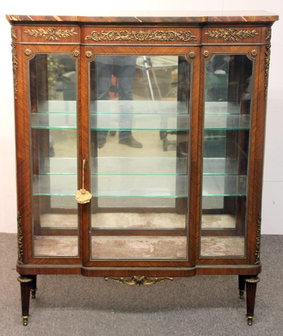 French Louis XVI-Style Marble-Top Vitrine: A French Louis XVI-style marble-top display vitrine cabinet in the style of F. Linke. The case with applied gilt bronze decoration depicting a central mask flanked by goats, as well as foliate design.