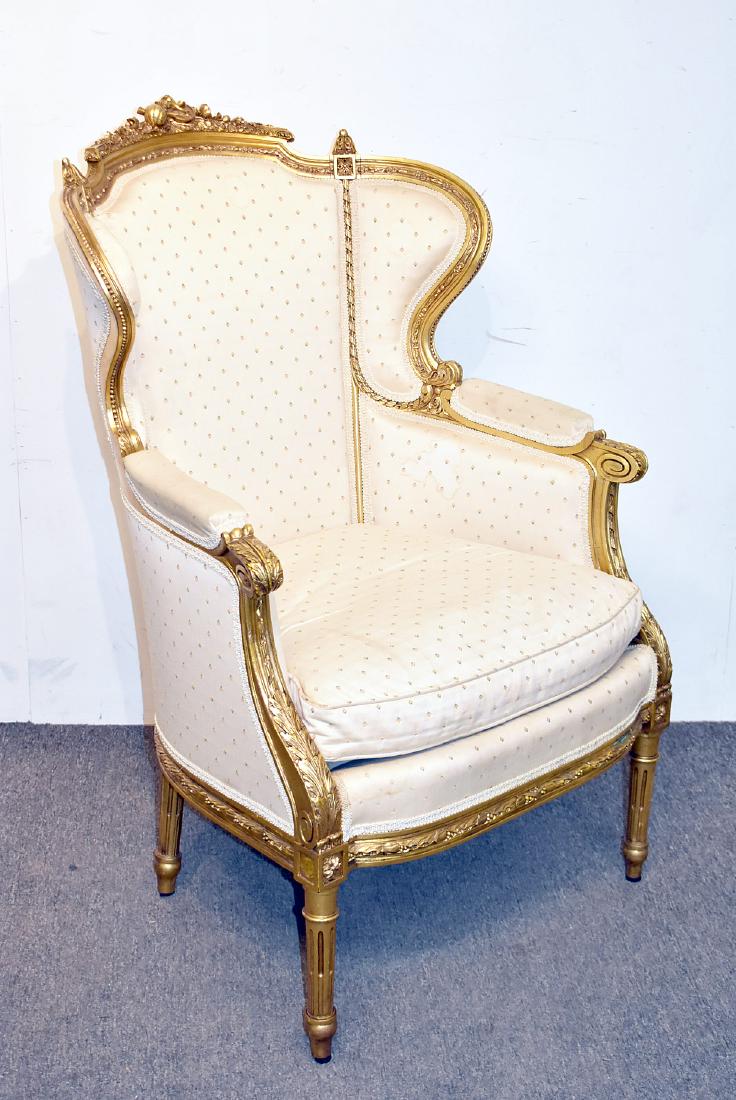 Louis XVI Giltwood Bergere: A Louis XVI giltwood upholstered bergère chair with foliate and musical instrument carved crest, raised on tapering, reeded legs. 43-1/2in tall x 29in wide x 25in deep.