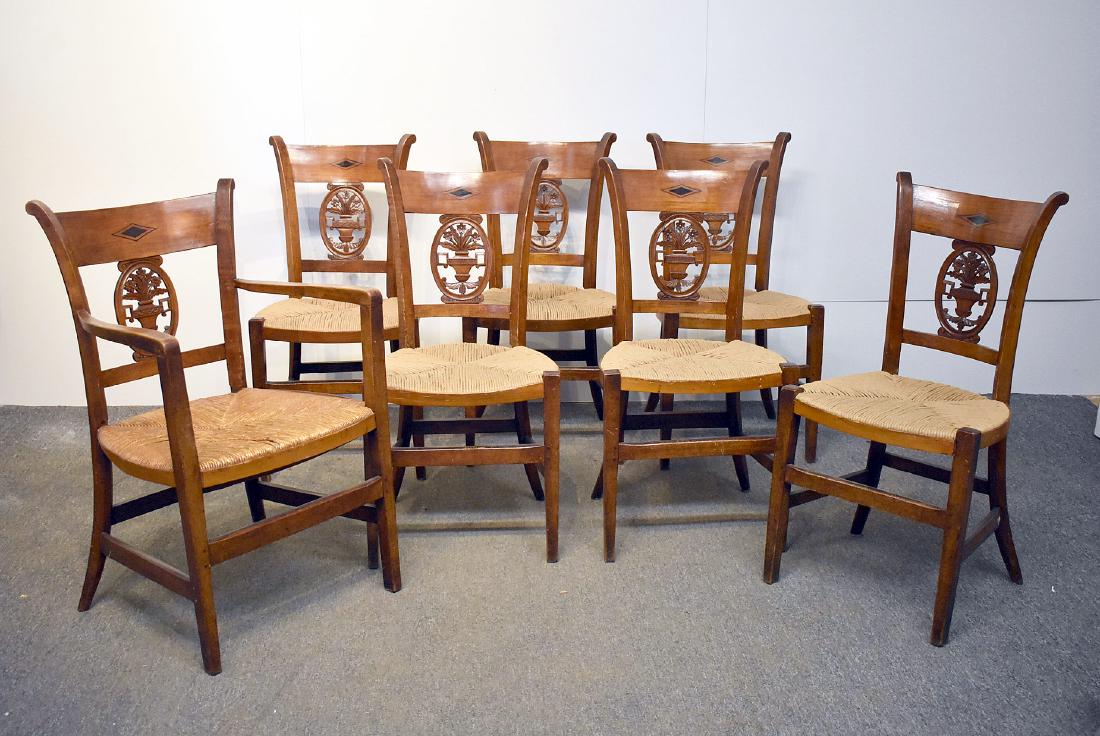 Seven French Provincial Carved Dining Chairs: A set of seven French Provincial carved dining chairs (one arm, 6 side) with ebony inlaid crest rails and floral basket splats over rush seats. Armchair: 35in tall x 21in wide x 18in deep.