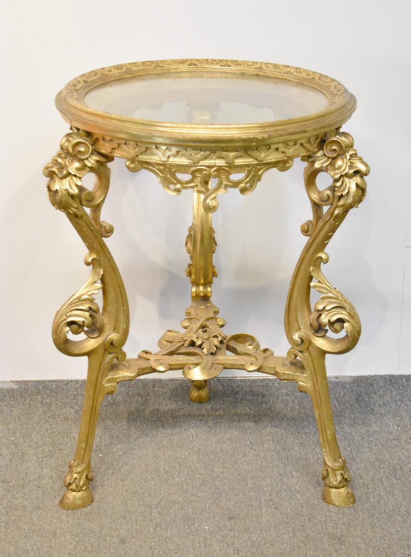 Louis XV-Style Giltwood Table: A Louis XV-style giltwood table, 19th / 20th Century, with an inset circular glass top over three acanthus-leaf carved cabriole legs with hoof feet, joined by a pierced stretcher. 31-1/2 in tall x 22 