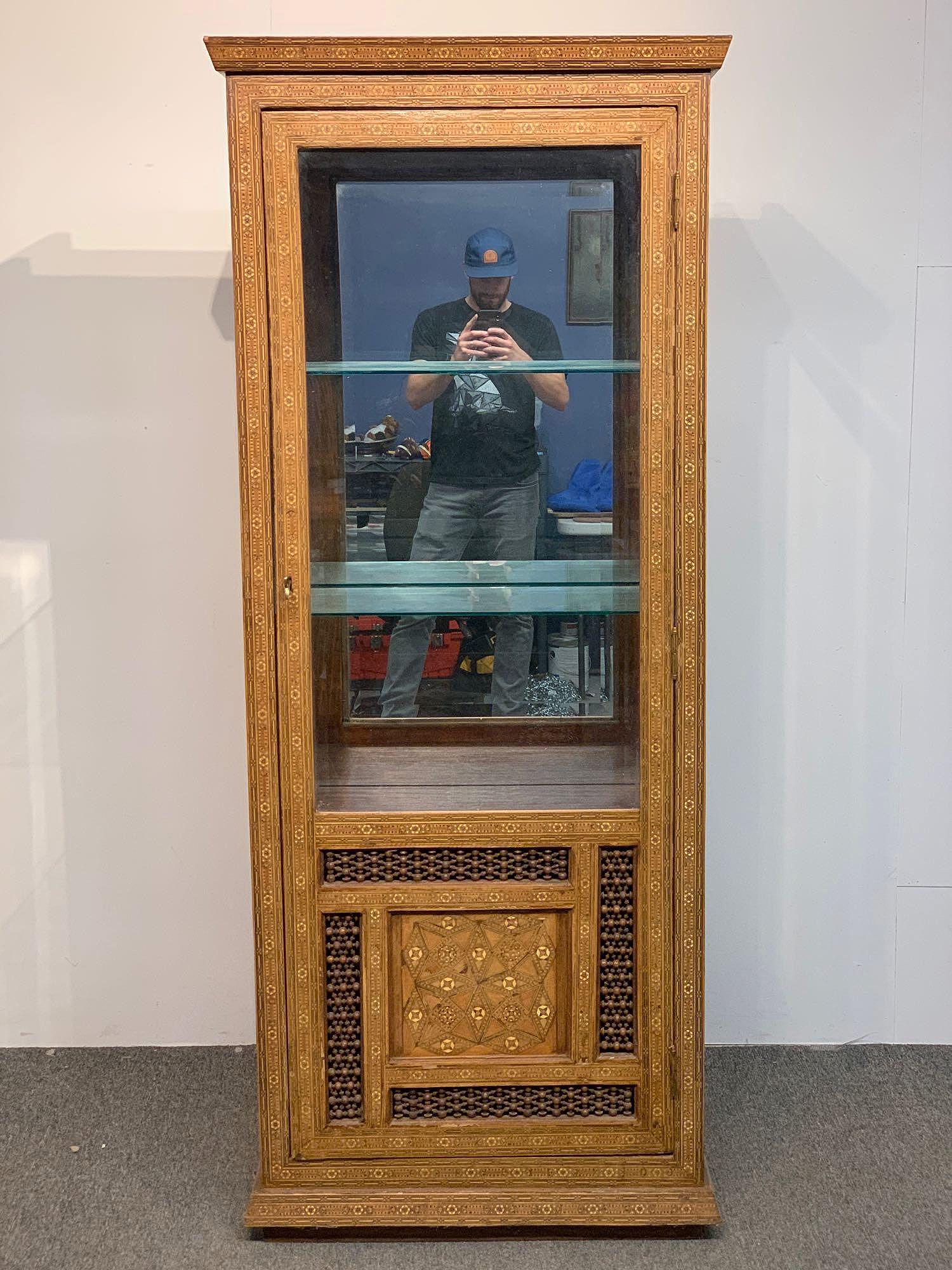 Syrian Glass Door Curio Cabinet: A Syrian glass door curio cabinet with mosaic inlaid and open baluster carved case, glass shelves, and mirrored back. 72in tall x 27-1/2in wide x 14in deep.