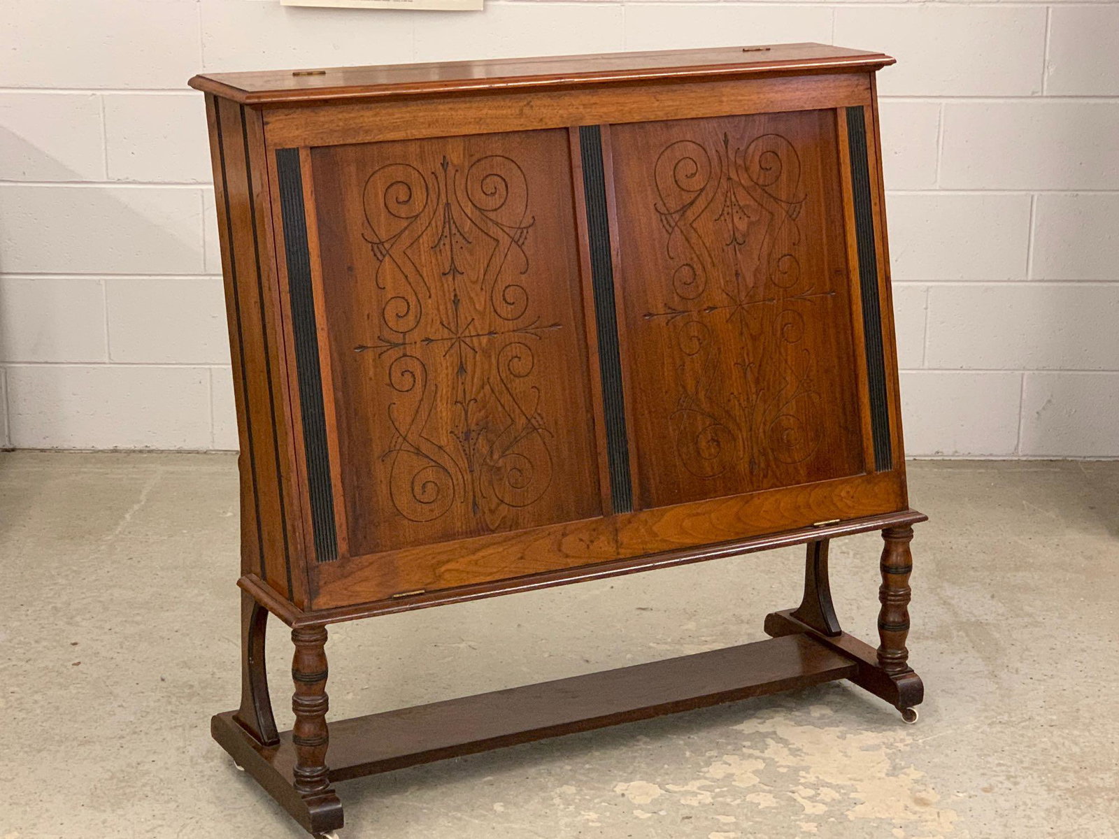 Victorian Carved Map Cabinet: A Victorian upright map or sheet music cabinet having a single hinged door with carved design flanked by ebonized reeded panels, raised on turned legs with casters. 43in tall x 43in wide x 16in deep.