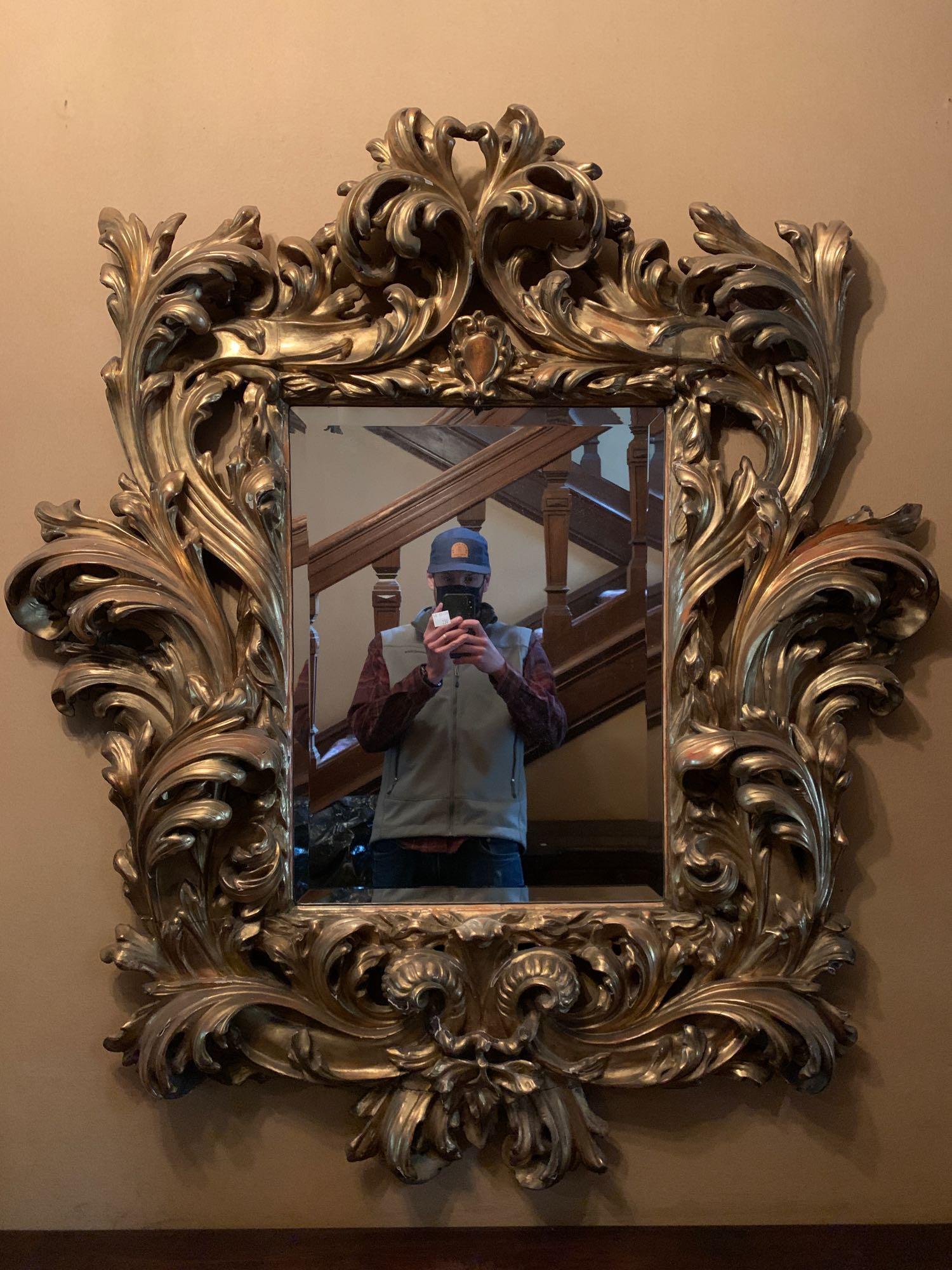 Italian Baroque Giltwood Mirror: An Italian baroque rococo giltwood wall mirror having a deeply carved and gesso frame, with beveled glass mirror plate. 50 in tall x 44 in wide.
