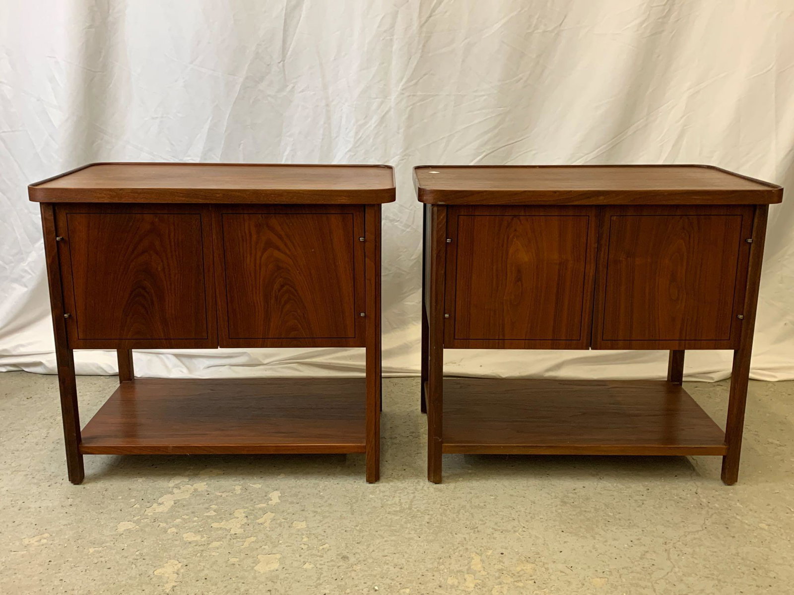 Pair of American Modern Walnut Night Stands: A pair of American midcentury modern walnut night stands having raised edges, two-door cabinets, and lower shelf stretchers. Unmarked. Each is 2 in tall x 26 in wide x 16 in deep.