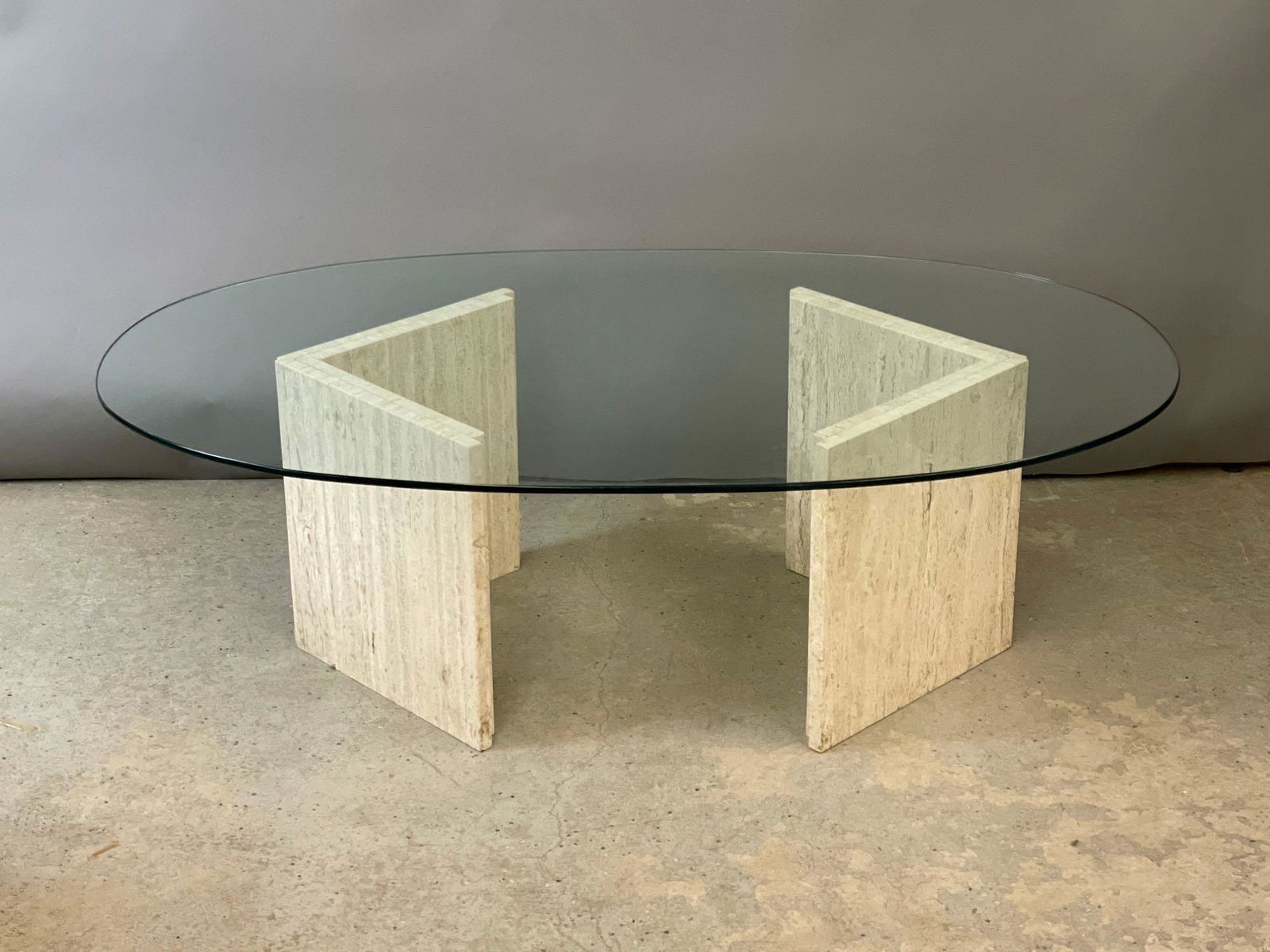Modernist Travertine and Glass Cocktail Table: A modernist cocktail table with an oval glass top over two travertine triangular bases. 15in tall x 50in wide x 30in deep.