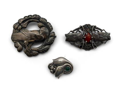 Three Georg Jensen Brooches: A collection of three Georg Jensen brooches to include a no. 70 sterling silver standing dove brooch, a no. 19 brooch with red gemstone cabochon (.830 silver), and a no. 100 brooch with green gemstone