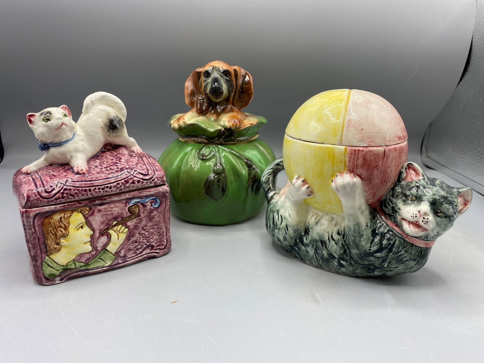 Three Figural Majolica Tobacco Jars: To include a cat with ball (5-1/2in tall, chips on lid); a cat on trunk (5-1/4in tall); a dog in sack (6in tall, chip on base). | Philadelphia Metro area delivery, NYC curbside delivery, and internati