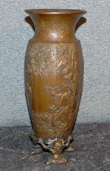 Chinese Bronze Vase: Unmarked bronze Chinese vase on figural base with three Buddhas with foo-dog heads. Embossed design with flowers, bee, a hut, and a Chinese junk. 10" tall; 4" diameter.