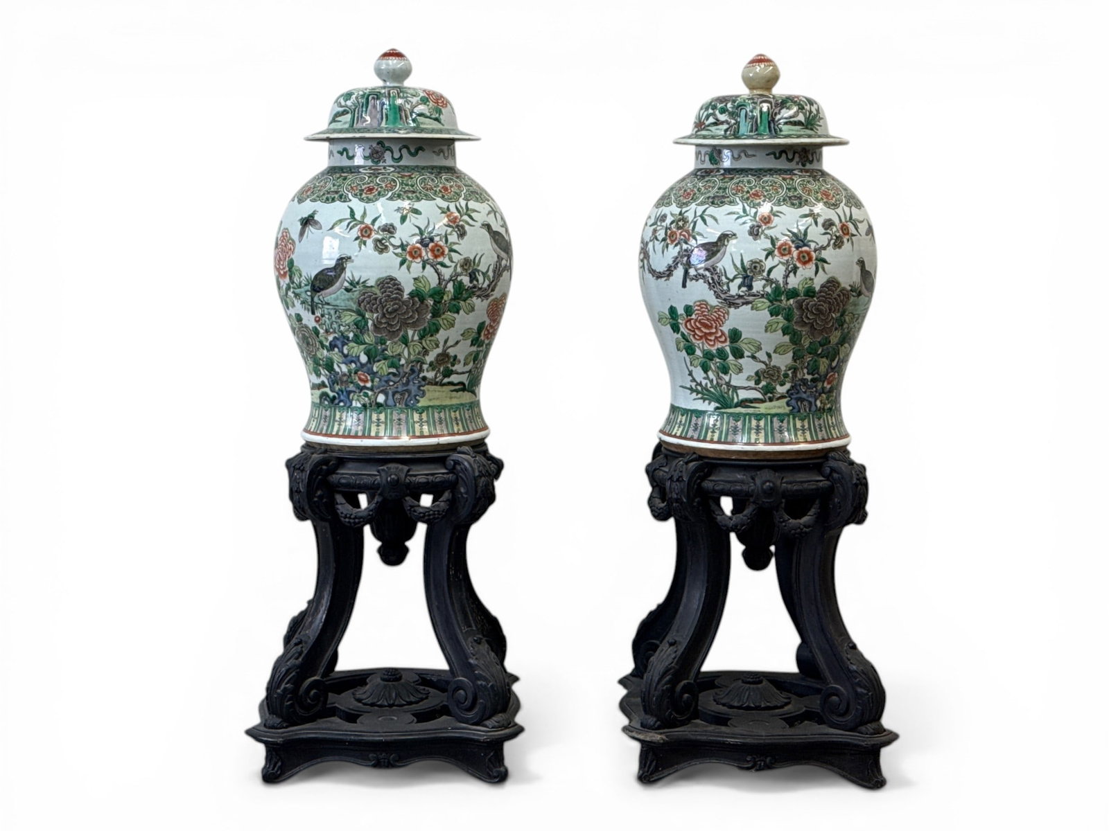 Pair of Chinese Export Famille Verte Porcelain Jars: A large pair of Chinese export famille verte porcelain covered jars decorated with enameled birds and verdant landscapes in vibrant colorations. Standing on raised carved wood display stands with swag