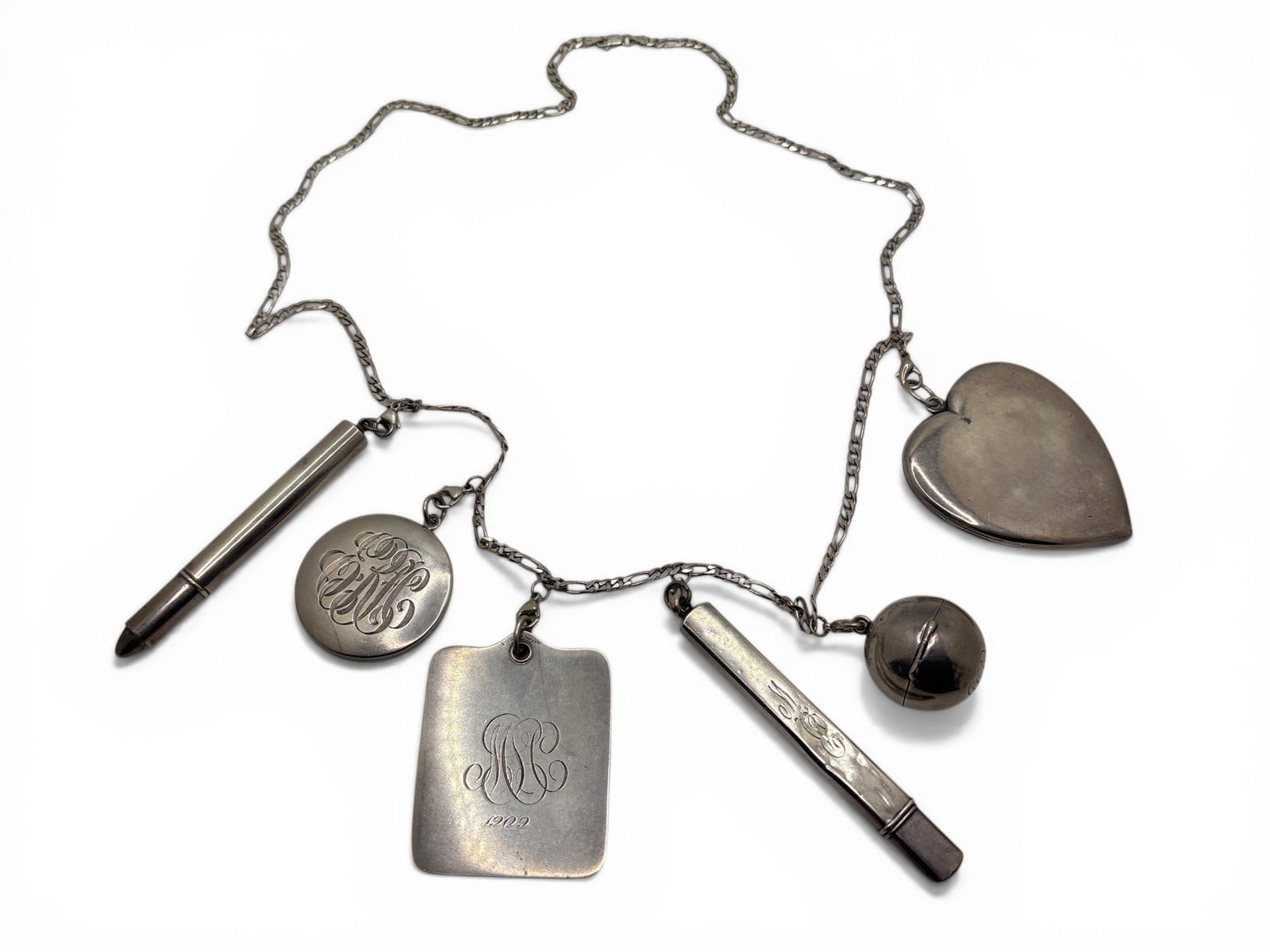 Sterling Figaro Chain Charm Necklace: A sterling figaro chain charm necklace, with six early 20th C charms consisting of a cylindrical vial, a monogrammed hand mirror, a monogrammed aide memoire engraved 1909, a monogrammed orb-form coin
