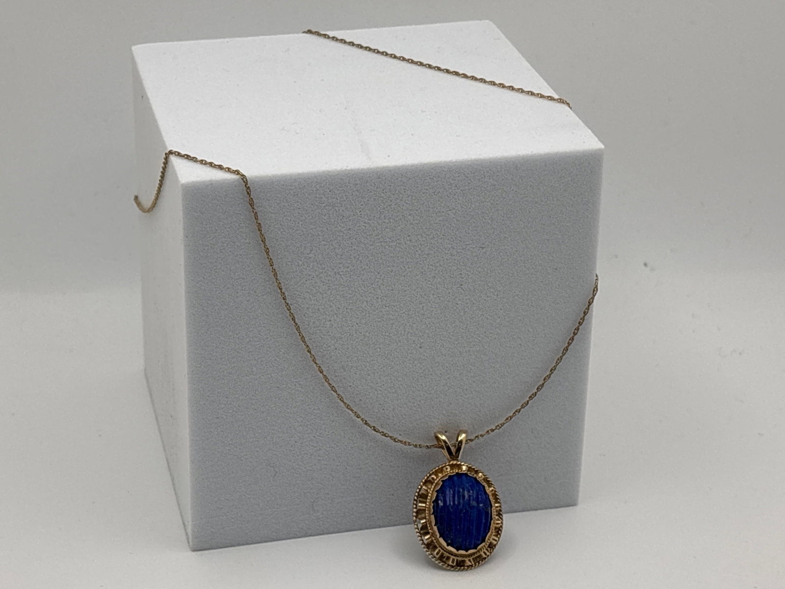 14K Yellow Gold Carved Lapis Pendant: A ribbed-carved 14mm x 10.4mm oval lapis cabochon in an undulating and twist-patterned 14K (tested) setting, suspended on a 14K (marked) yellow gold chain. 14-1/2in long. Pendant: 1-1/8in tall x