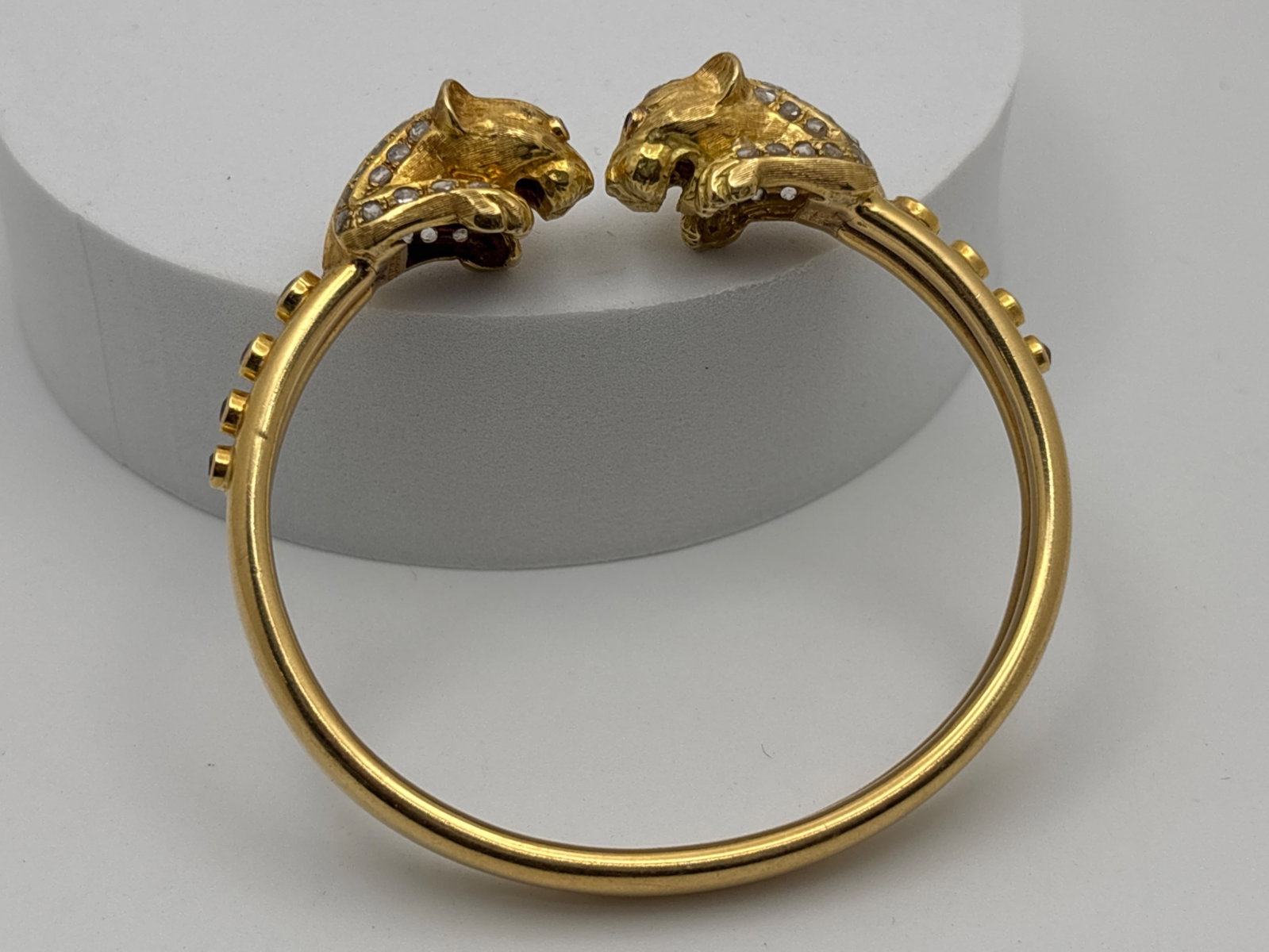 18K Illias Lalaounis Leopard Head Bracelet: An open bangle with two facing leopard heads, each with ruby eyes and diamond accents, the bracelet set with 1.4mm - 2.6mm sapphires and rubies. Maker's mark stamp in the interior: K18. 2-3/8in inner