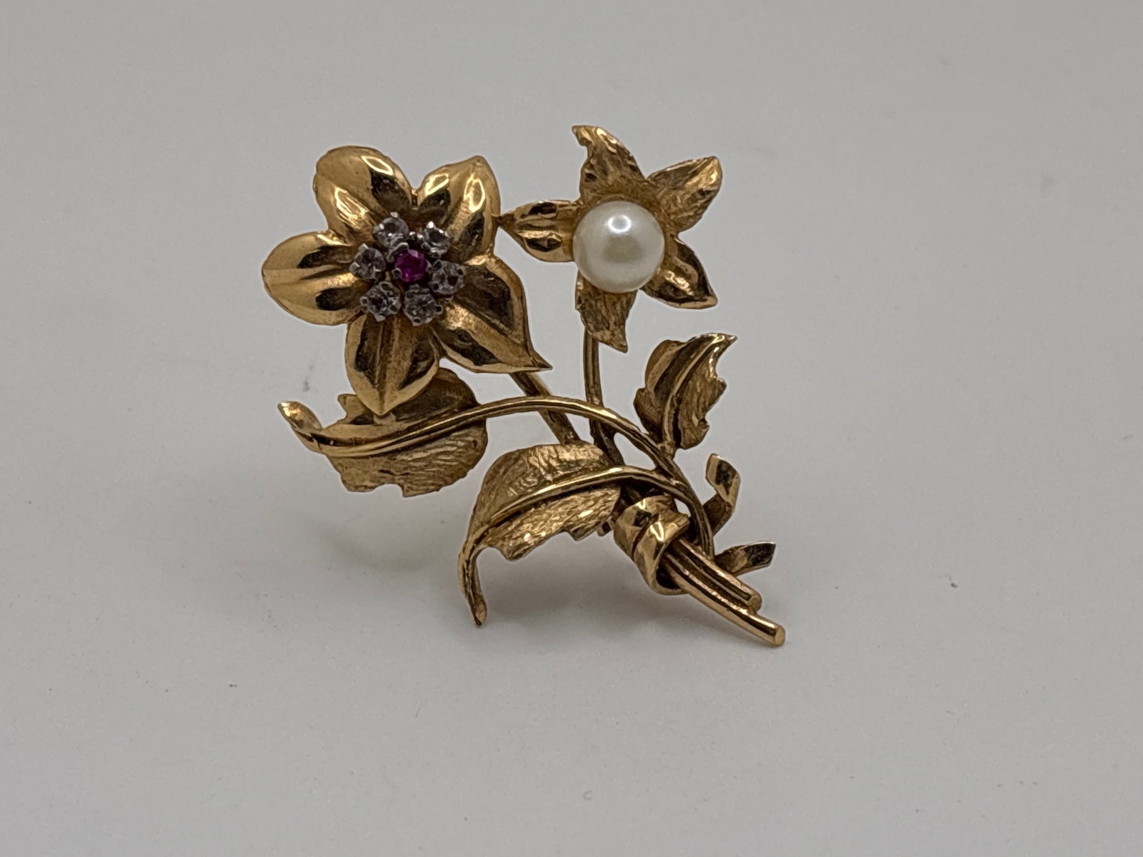 18K Ruby and Pearl Floral Brooch: An 18K yellow gold brooch of blooming flowers, the center of the largest bloom set with a 2.1mm ruby in a six 2.4mm diamond surround. The smaller bloom has a 6.4mm pearl at center, 1-7/8in tall x 1-1/