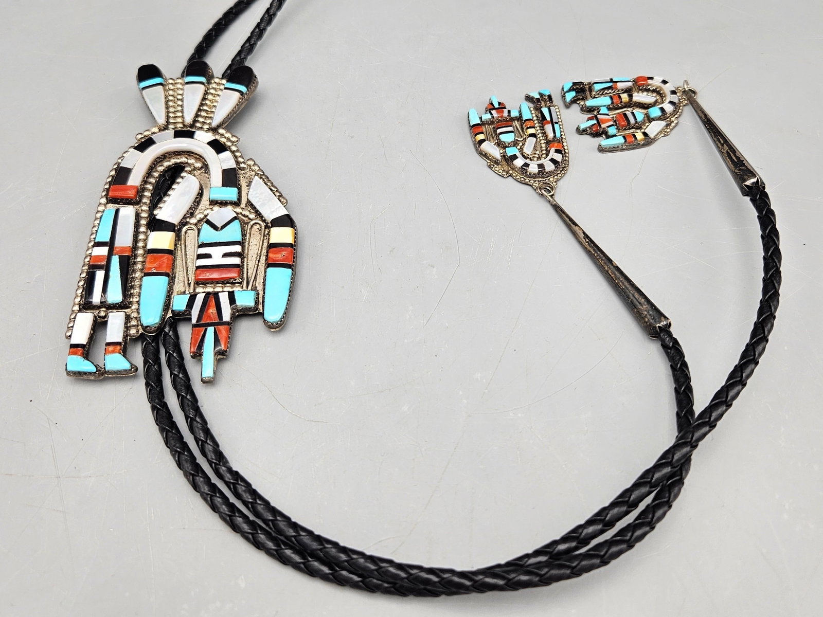 Zuni Silver Rainbow Dancer Bolo Tie: A Zuni silver rainbow dancer figural bolo tie with smaller rainbow dance tie caps, set with turquoise, onyx, coral and mother of pearl. Signed. 20-1/2in long. Figure: 4in tall x 2-1/2in wide. 52.86 dw