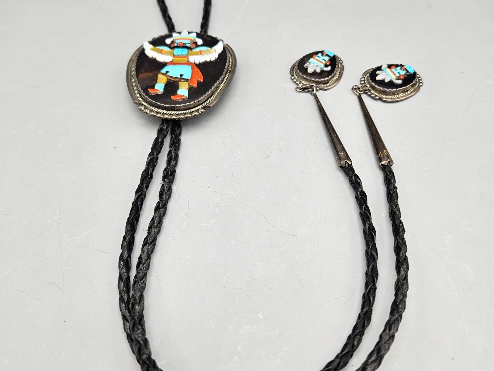 Beverly Estate Zuni Dancer Bolo Tie: A Beverly Estate (Zuni, 20th C.) Zuni silver bolo tie with eagle dancer figure slide and tie cap set with turquoise, onyx, mother of pearl and coral. Signed. 20in long. Slide: 2in tall x 3in wide. 45.