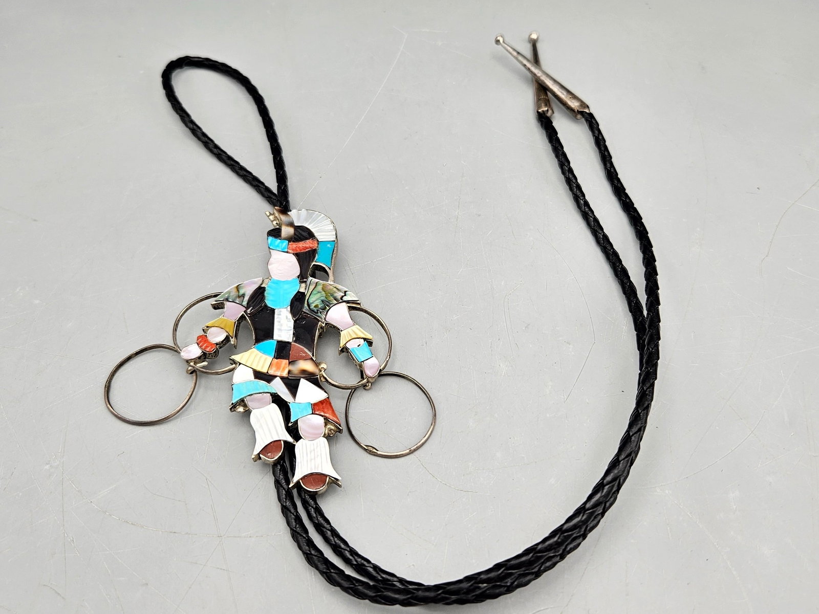 Jonathan Beyuka Zuni Silver Dancer Bolo: Jonathan Beyuka (Zuni, 20th C.) A silver bolo tie with Zuni ceremonial dancing figure with inlaid turquoise, onyx, and mother of pearl. Signed. 18in long. Figure: 3-1/2in tall x 3in wide. 27.52 dwt. |