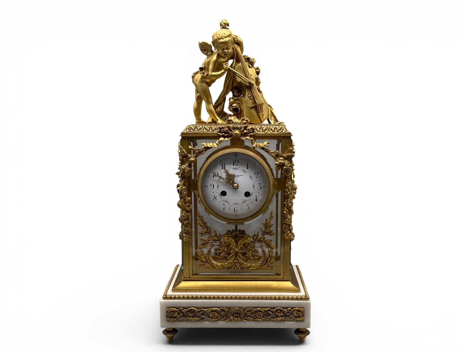 French Louis XVI-style Bronze and Crystal Mantel Clock: A Harris and Harrington French Louis XVI-style bronze mantel clock having a crystal and bronze case with foliage swag and a cherub playing a cello finial, resting on a marble and bronze plinth. Signed